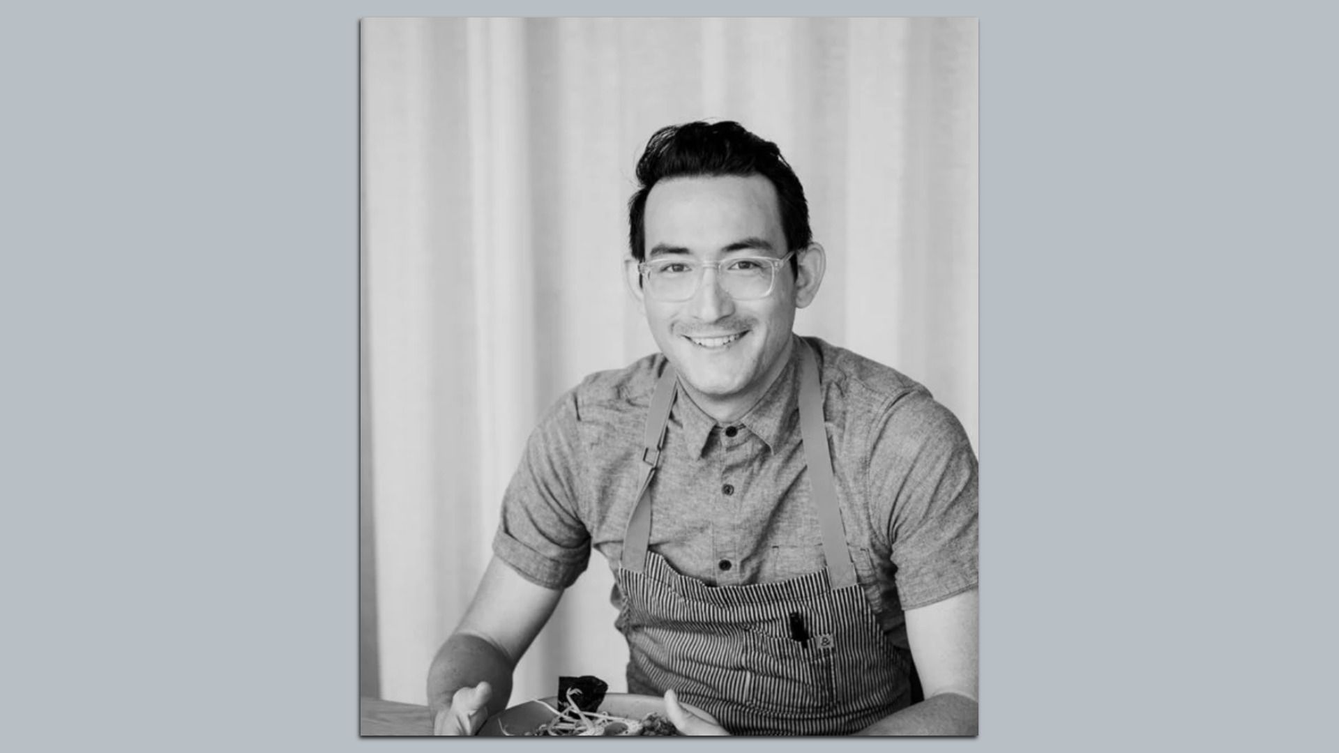 Black-and-white portrait of a smiling man with glasses wearing a striped apron, seated at a table with food, in front of light curtains.