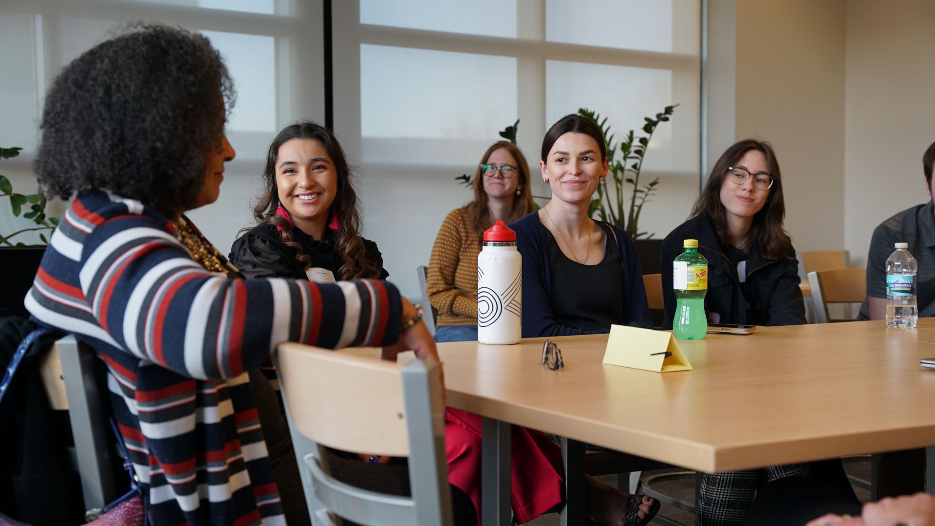 Five people sit around a light wood table in a bright room with large windows and plants. They smile and chat. On the table: a white bottle with red cap, a green bottle, a water bottle, and a yellow card.