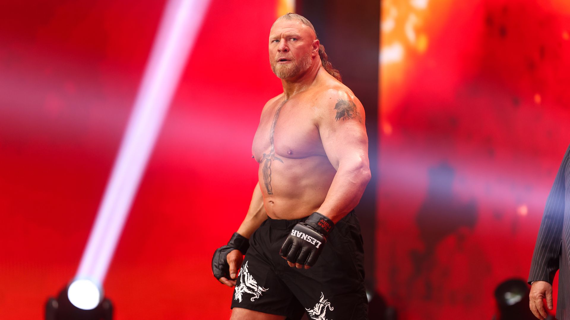 Shirtless, muscular man with blond beard and ponytail on a red-lit stage, wearing black gloves and shorts with white designs; tattoos on chest and arm, he looks surprised.