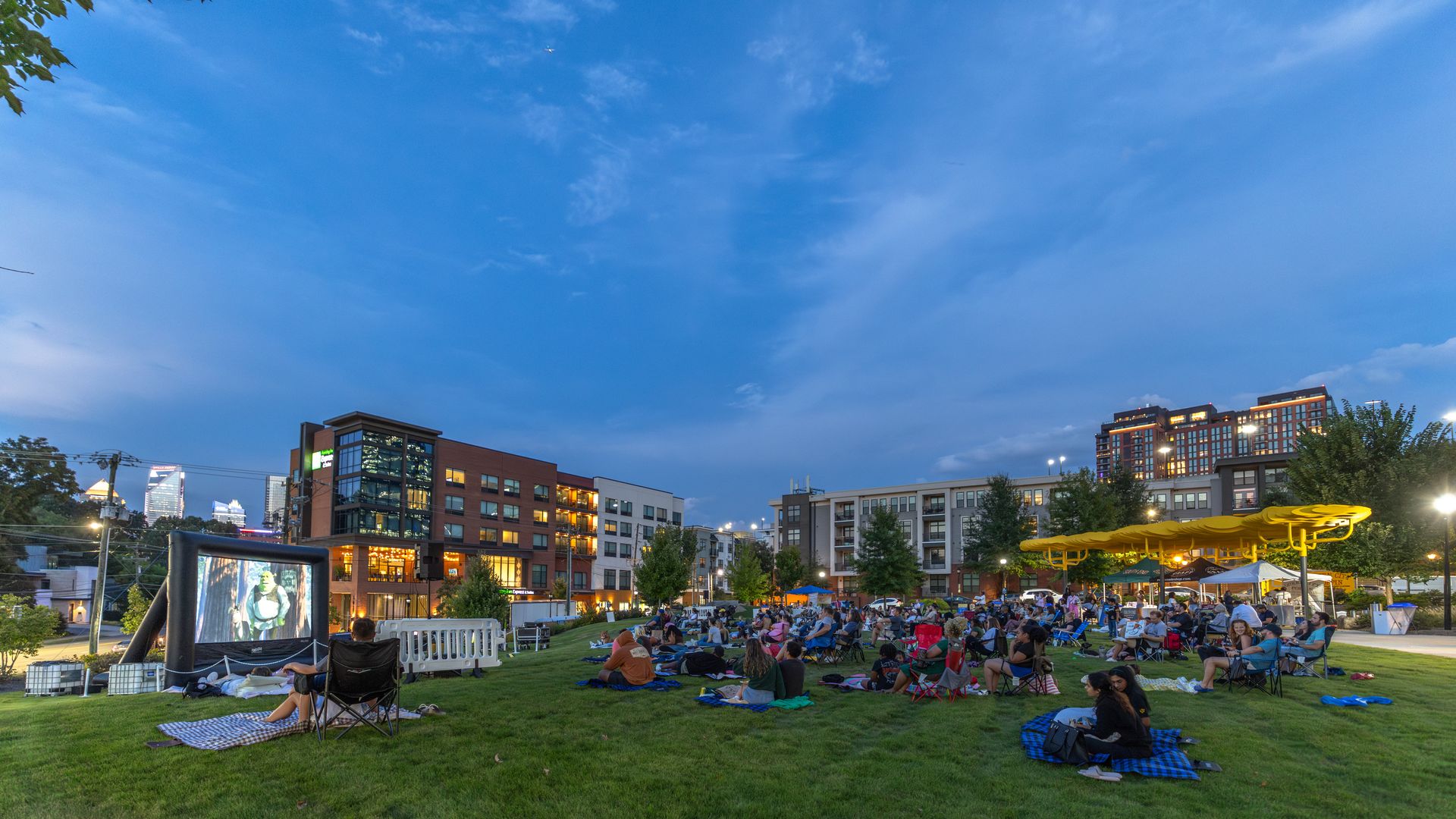 Outdoor movie night in a city park at dusk. People sit on blankets and chairs facing a large screen; yellow umbrellas at right, modern buildings in the background under a blue evening sky.