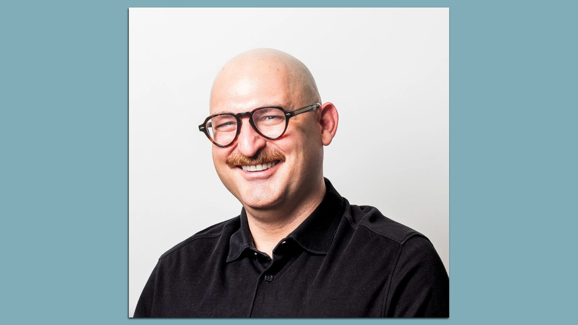 Smiling man with round glasses and a mustache, wearing a black shirt, posed against a light gray backdrop with a teal border.