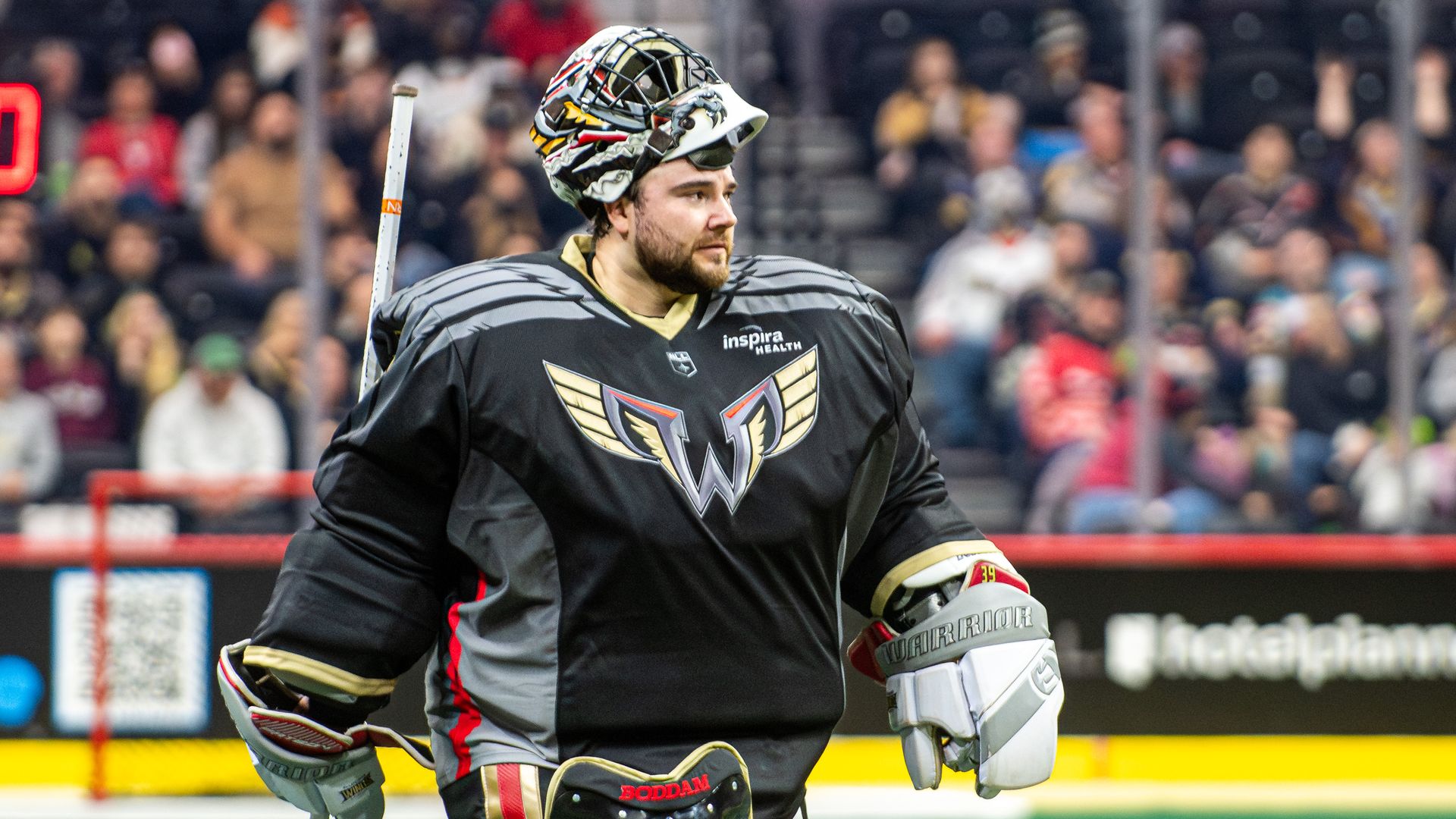 Lacrosse goalie in a black jersey with a gold wing emblem stands on a green field, wearing pads and a helmet on his head, holding a stick near the goal as a crowd watches.