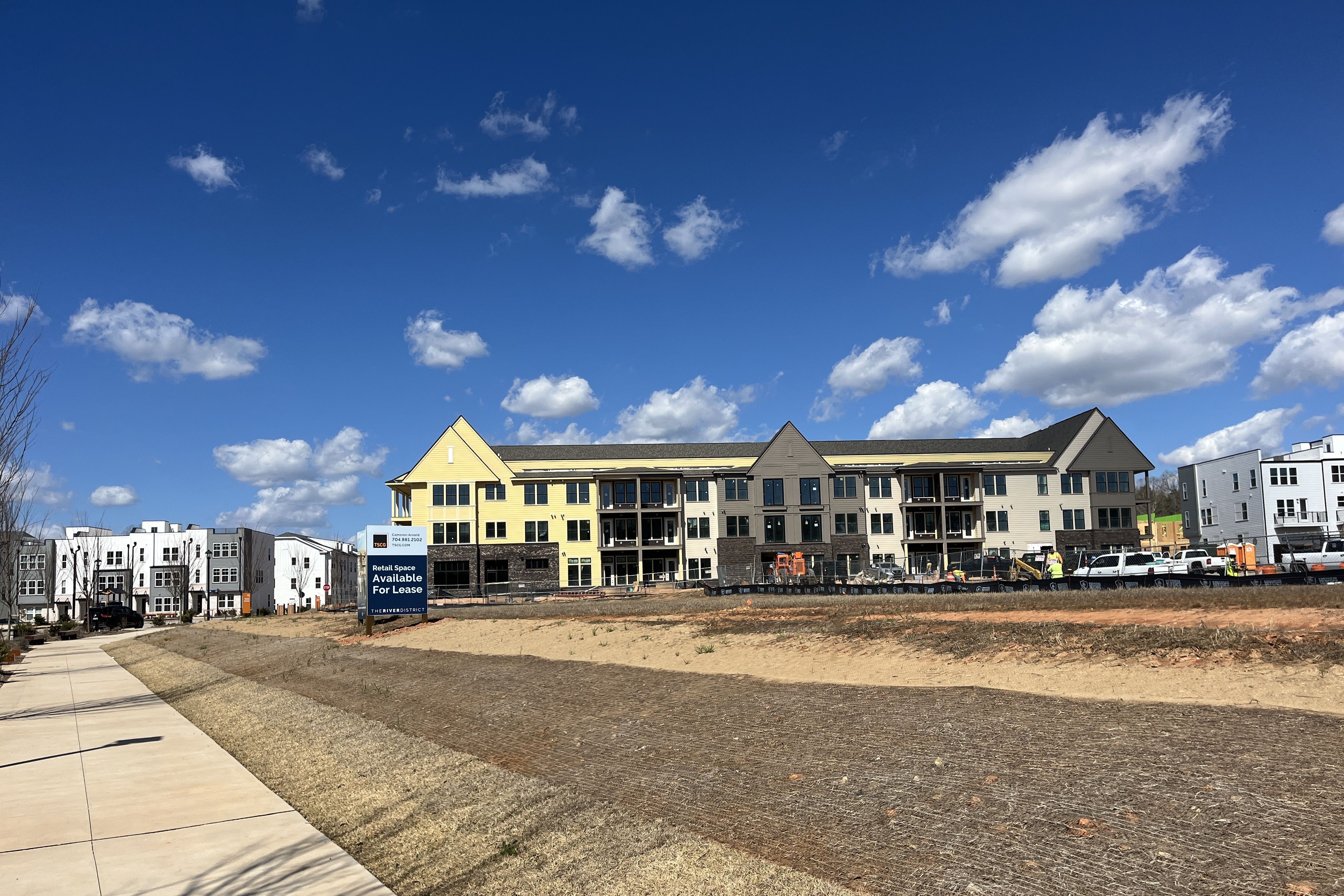 Row of modern beige and yellow apartment buildings under a bright blue sky with white clouds; a sign reads "Retail Space Available For Lease" near a dirt lot and sidewalk.