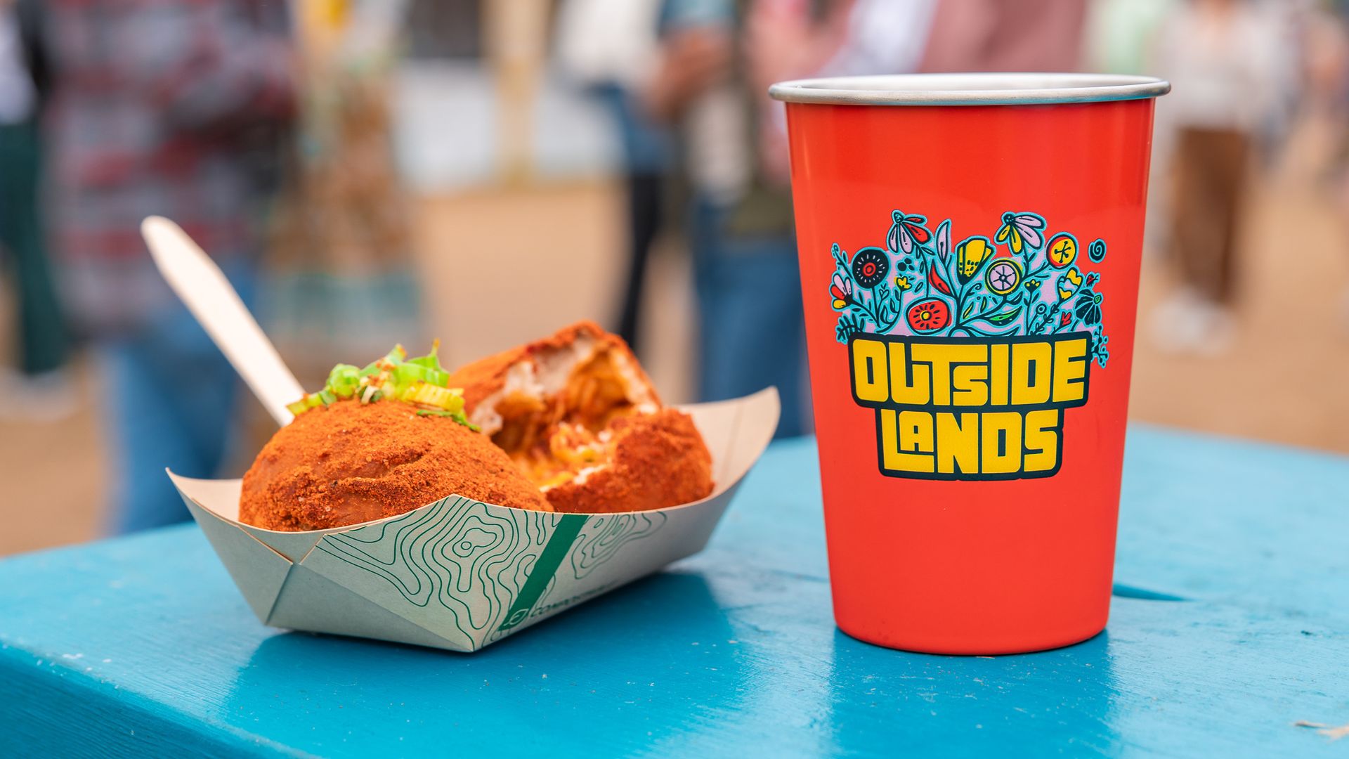 Red cup with a blue floral design and the text "OUTSIDE LANDS" sits on a blue table; in front is a fried-food tray, while a blurred crowd is in the background.