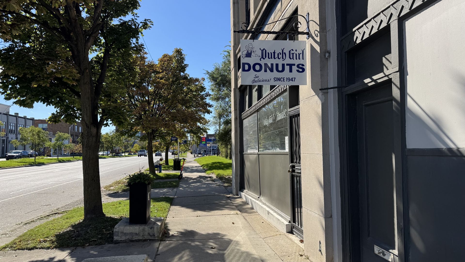 Sunlit street scene: gray building with a hanging sign reading "Dutch Girl DONUTS" in blue, "Since 1947". Trees line the sidewalk; distant cars and a person near the curb.