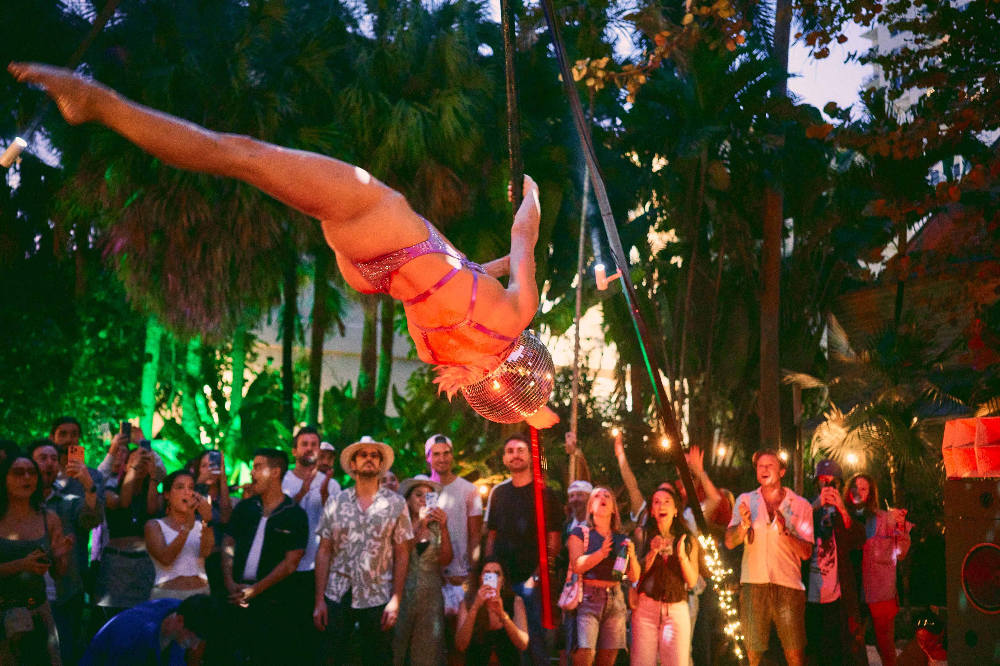 A performer in a purple costume and disco ball helmet hangs upside down from a pole, entertaining a crowd at an outdoor nighttime event surrounded by palm trees and colorful lights.
