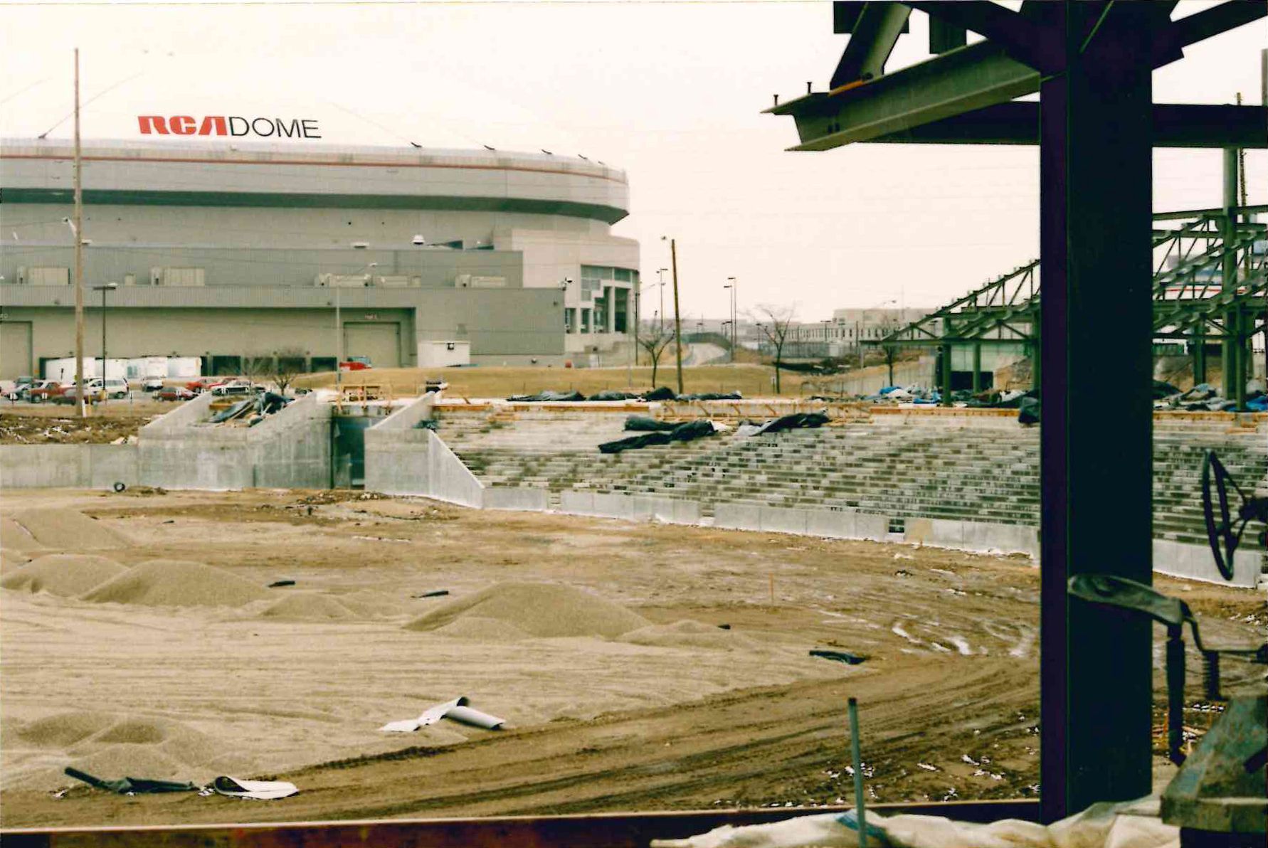 Wide view of a construction site with the rounded RCA Dome on the left, labeled "RCA DOME". Foreground shows concrete bleachers, dirt, and debris; right side features dark steel framework of unfinished structures.
