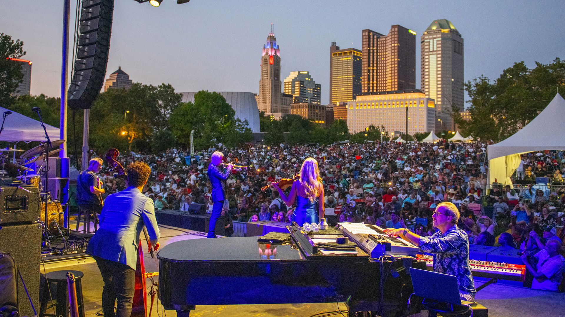 Outdoor concert at dusk with a large crowd, musicians on stage (piano and sax), a tall speaker stack on the left, and a brightly lit city skyline in the background.