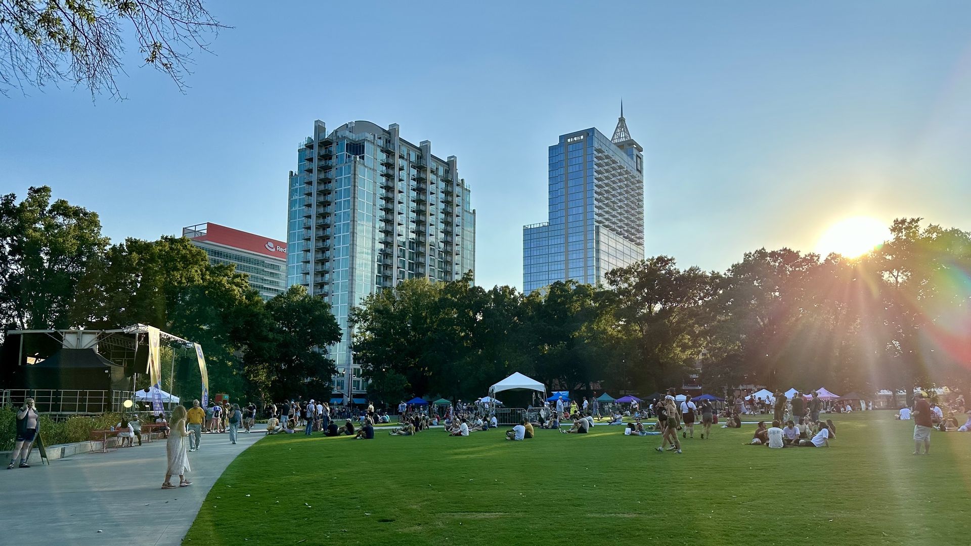 Sunlit urban park with a grassy field and groups of people relaxing; modern glass skyscrapers rise in the background under a clear blue sky, with the sun on the right creating lens flare.