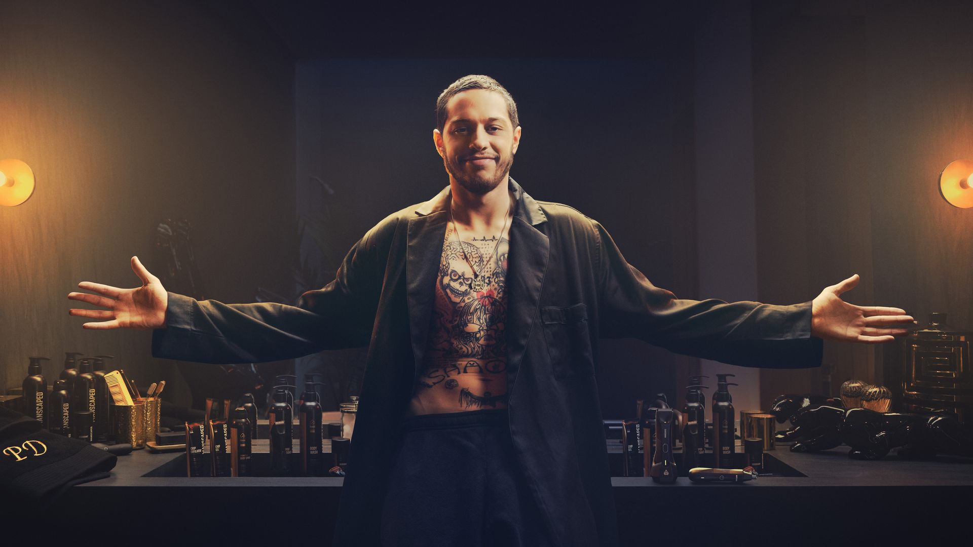 Comedian Pete Davidson stands in a darkened room wearing a black blazer with his hands extended outwards.