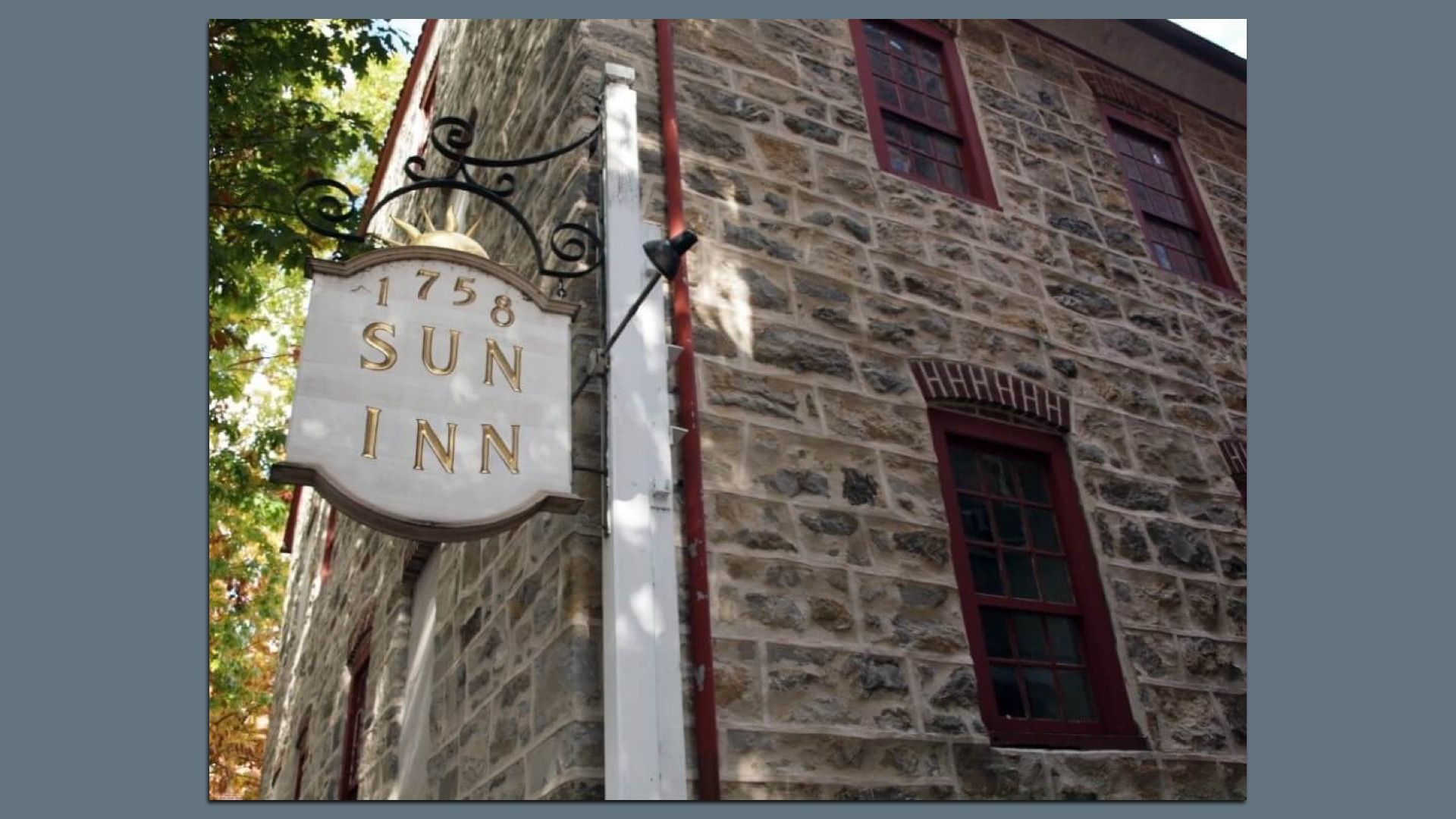 Stone inn exterior with a white sign reading "1758 SUN INN" in gold letters, hanging from a decorative black bracket. Red-trimmed windows, white post, and leafy trees frame the scene.