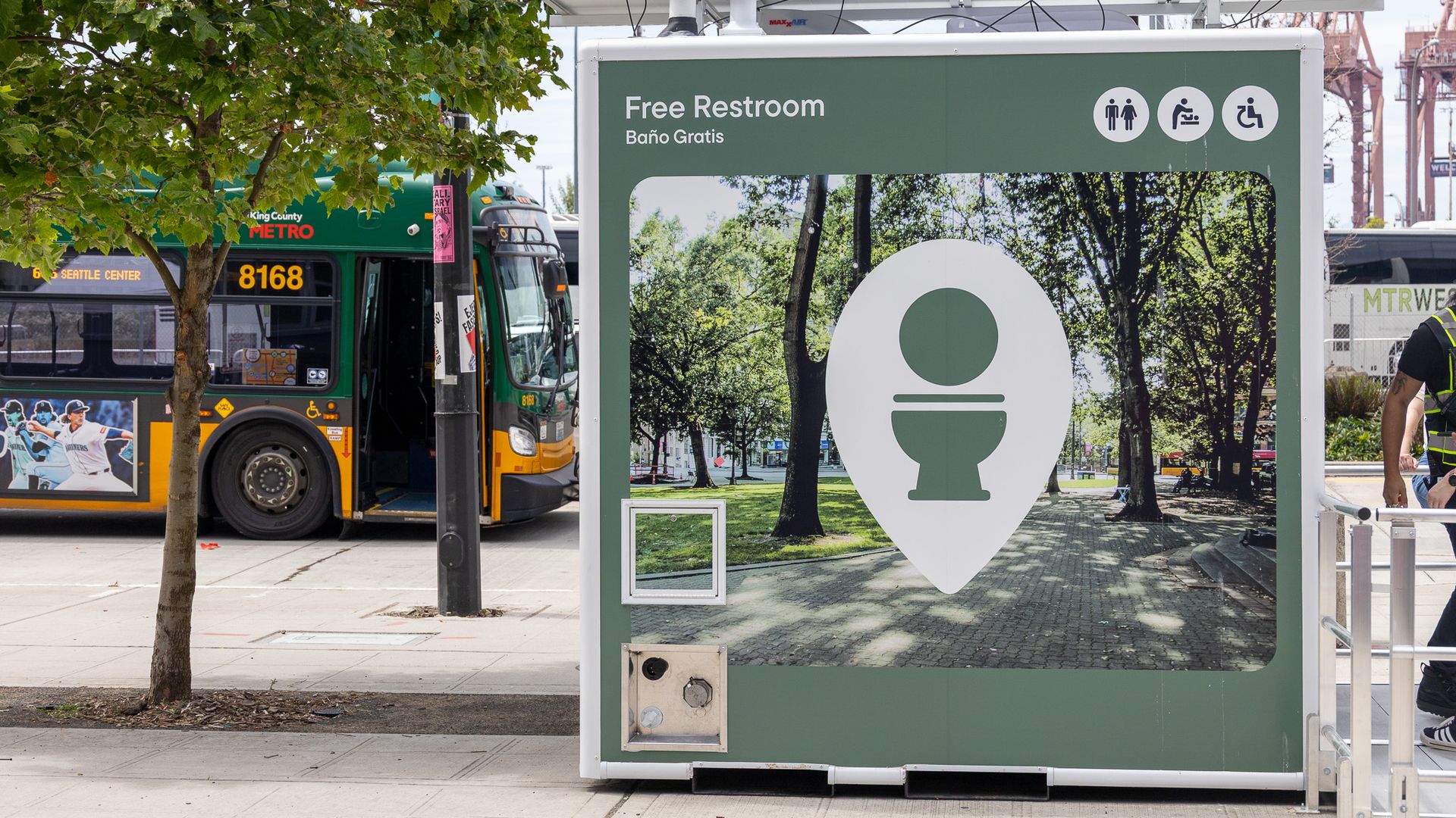 Large green signboard reads Free Restroom Baño Gratis with accessibility icons; a King County Metro bus is on the left, with trees in the foreground and construction cranes in the distance.