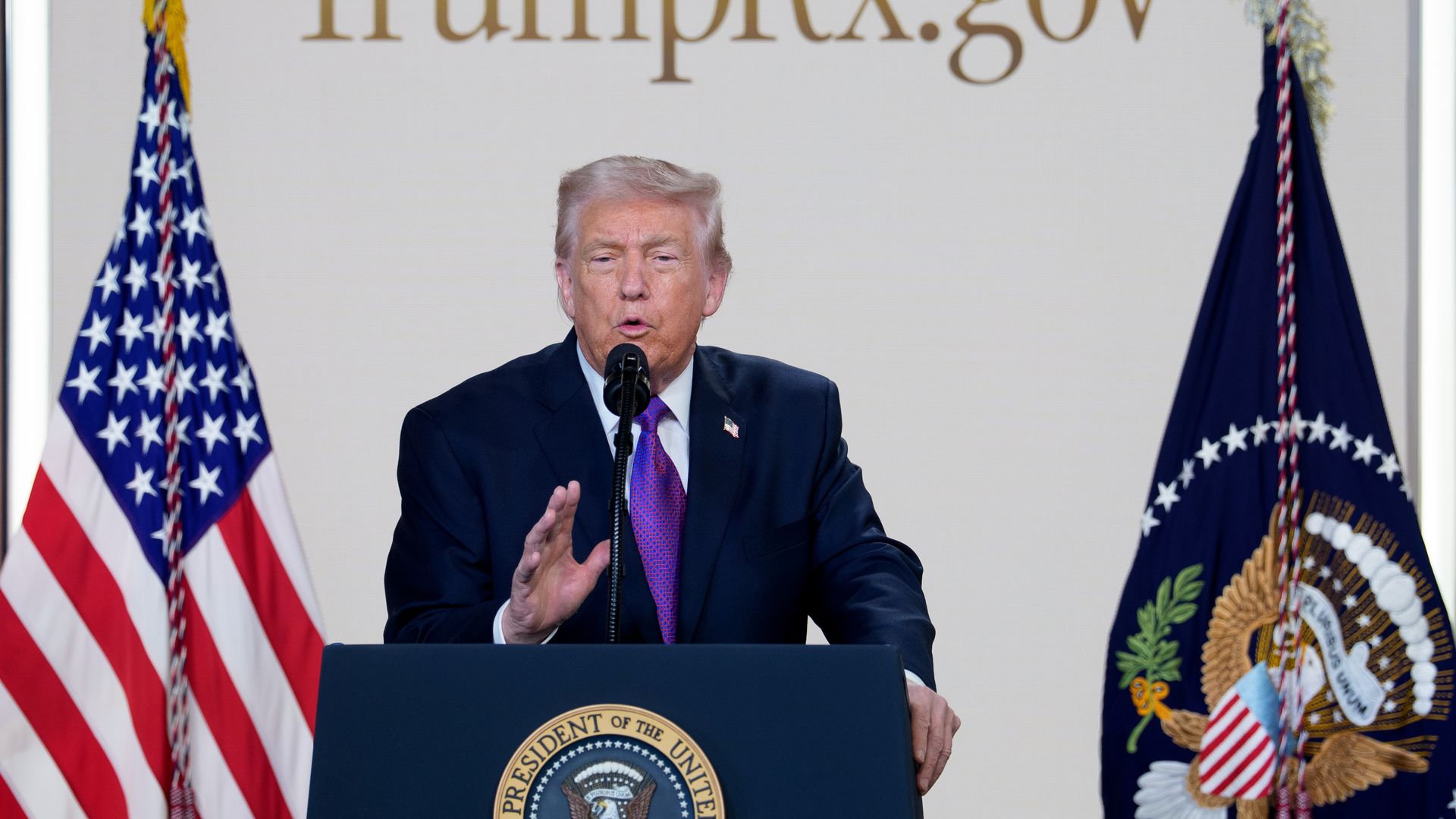 Donald Trump speaking at a podium with the presidential seal, flanked by the U.S. flag and presidential flag, with text "TrumpRx.gov" displayed behind him.