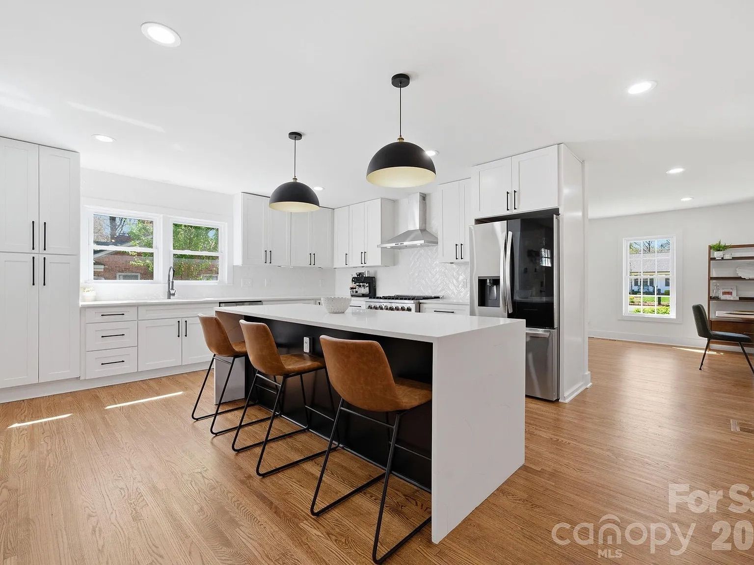 Bright modern kitchen with white cabinets, large white island, brown leather stools, stainless steel fridge and range, black pendant lights, wood floor, window view of greenery.