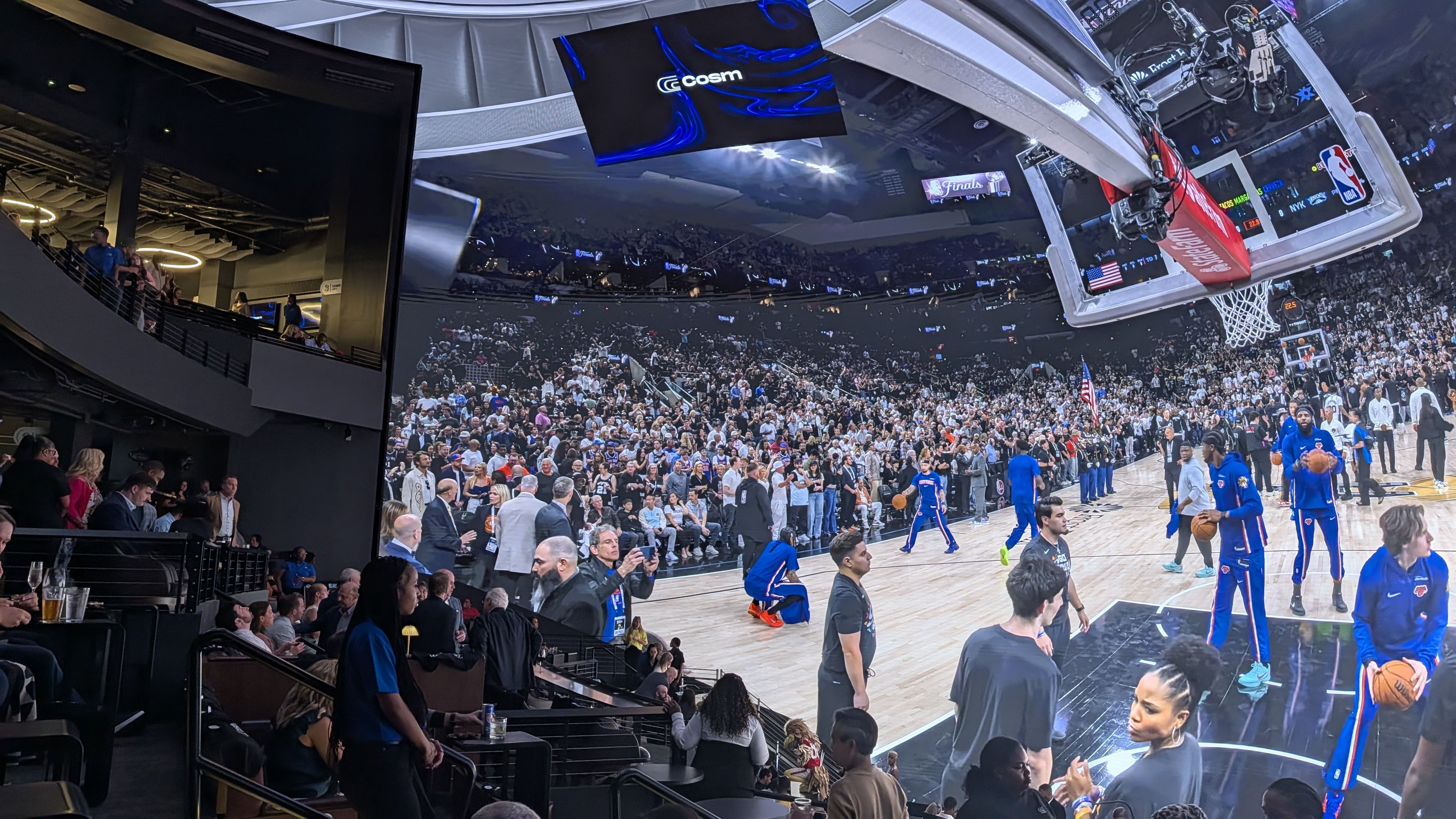 Guests watch a live view of a basketball team warming up inside an Indoor arena filled with a large crowd; a hoop and backboard loom on the right while blue-clad players warm up on the court and fans fill the stands.