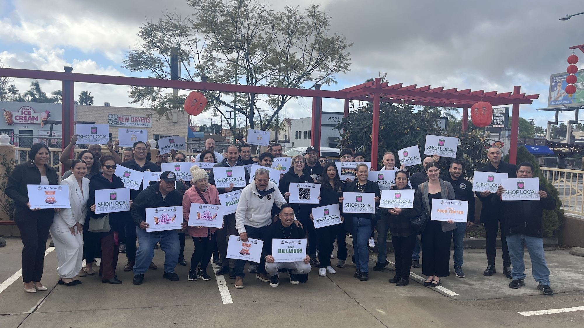 Business owners pose for a photo outside holding signs with shop names and "shop local City Heights."