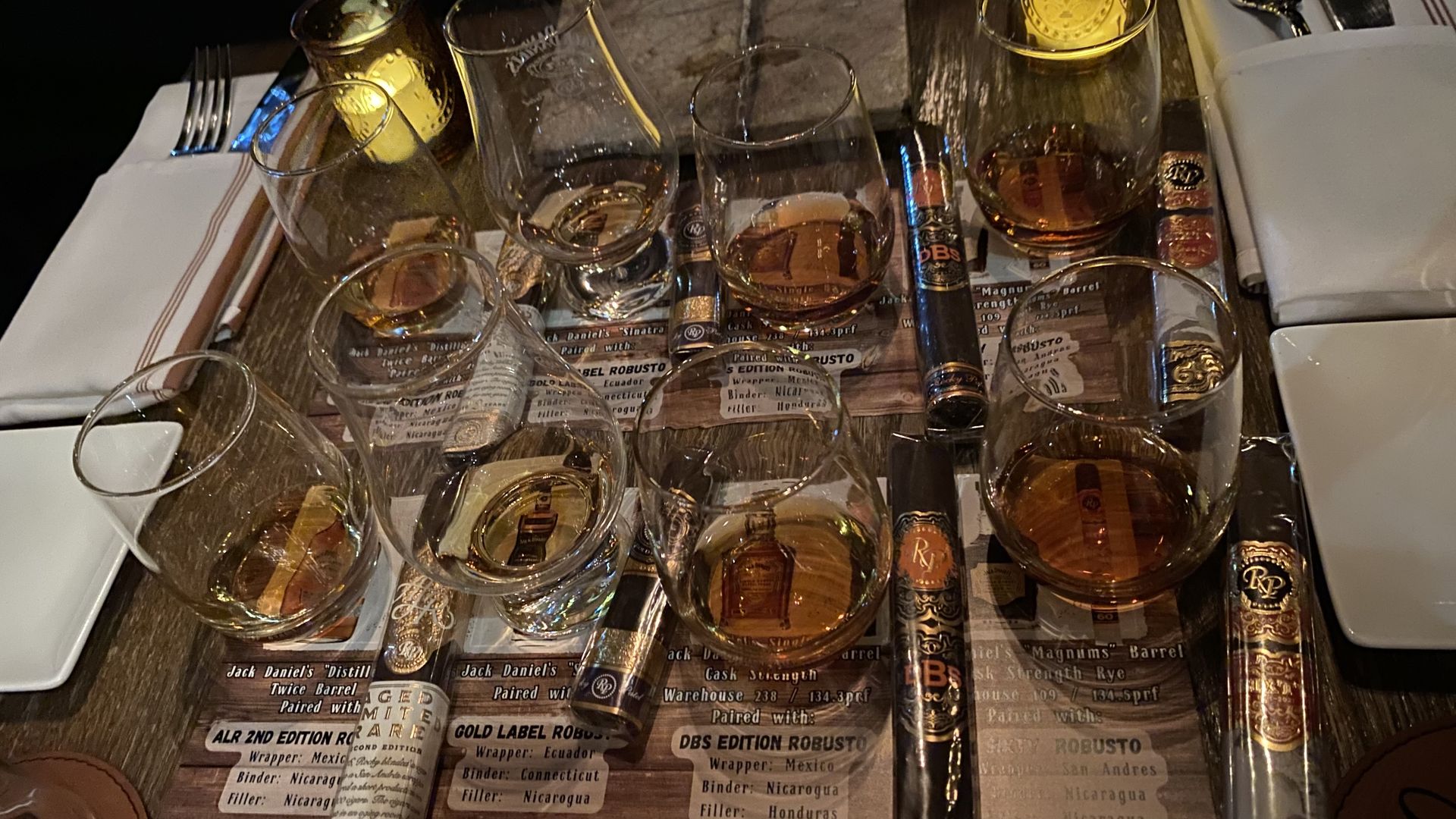 Eight small glasses with small amounts of whiskey in them, with a cigar next to each one. 
