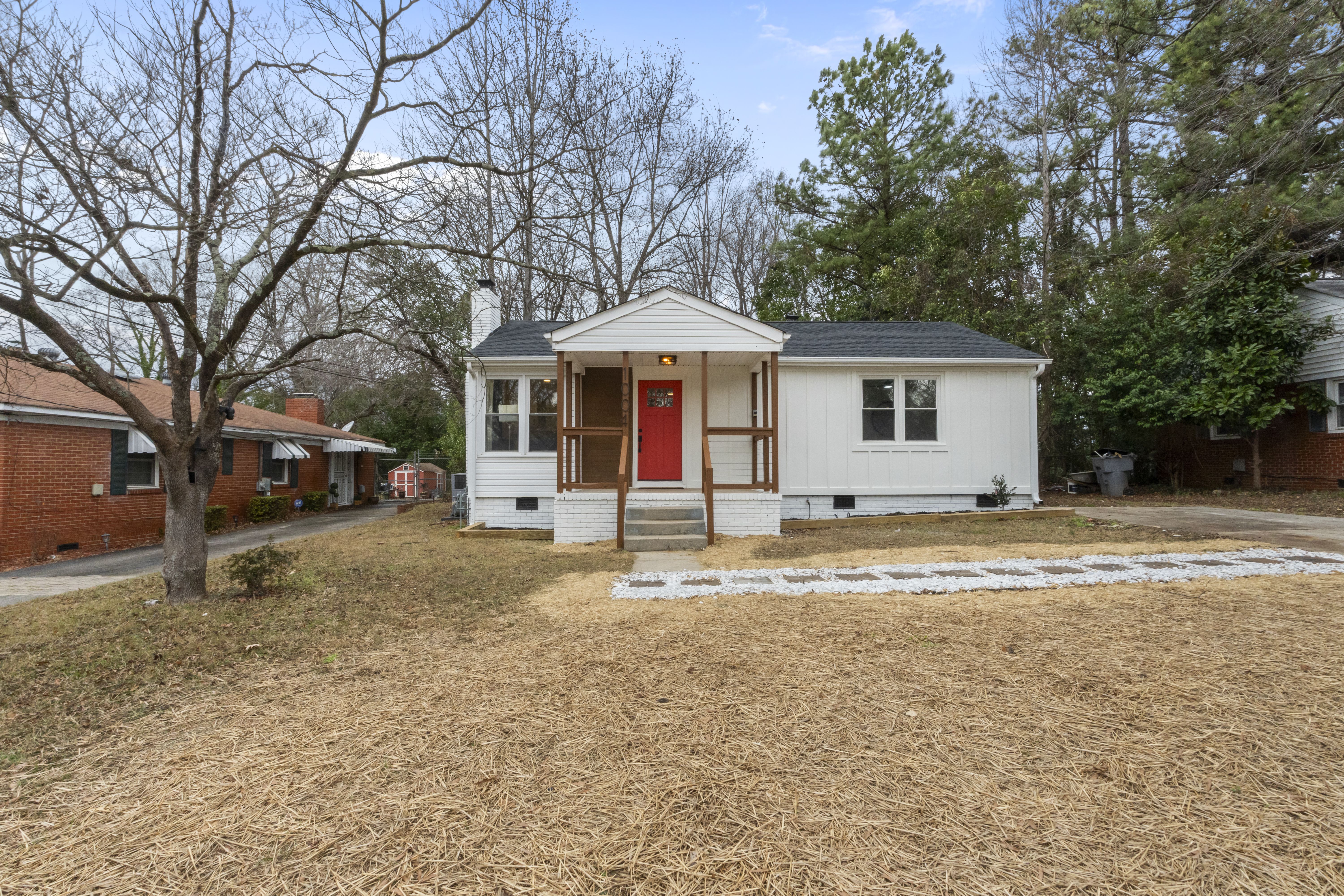Small white house with dark roof and red front door centered on plot with mulch-covered yard, leafless trees behind and nearby brick houses on each side under blue sky.