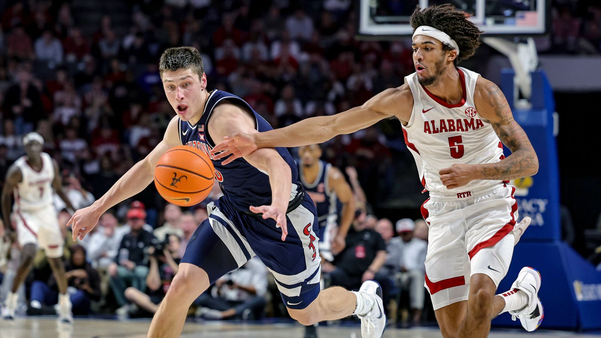 Two college basketball players in action: one in navy blue uniform reaching for a ball, the other in white Alabama uniform #5 trying to block him on a court with spectators in background.