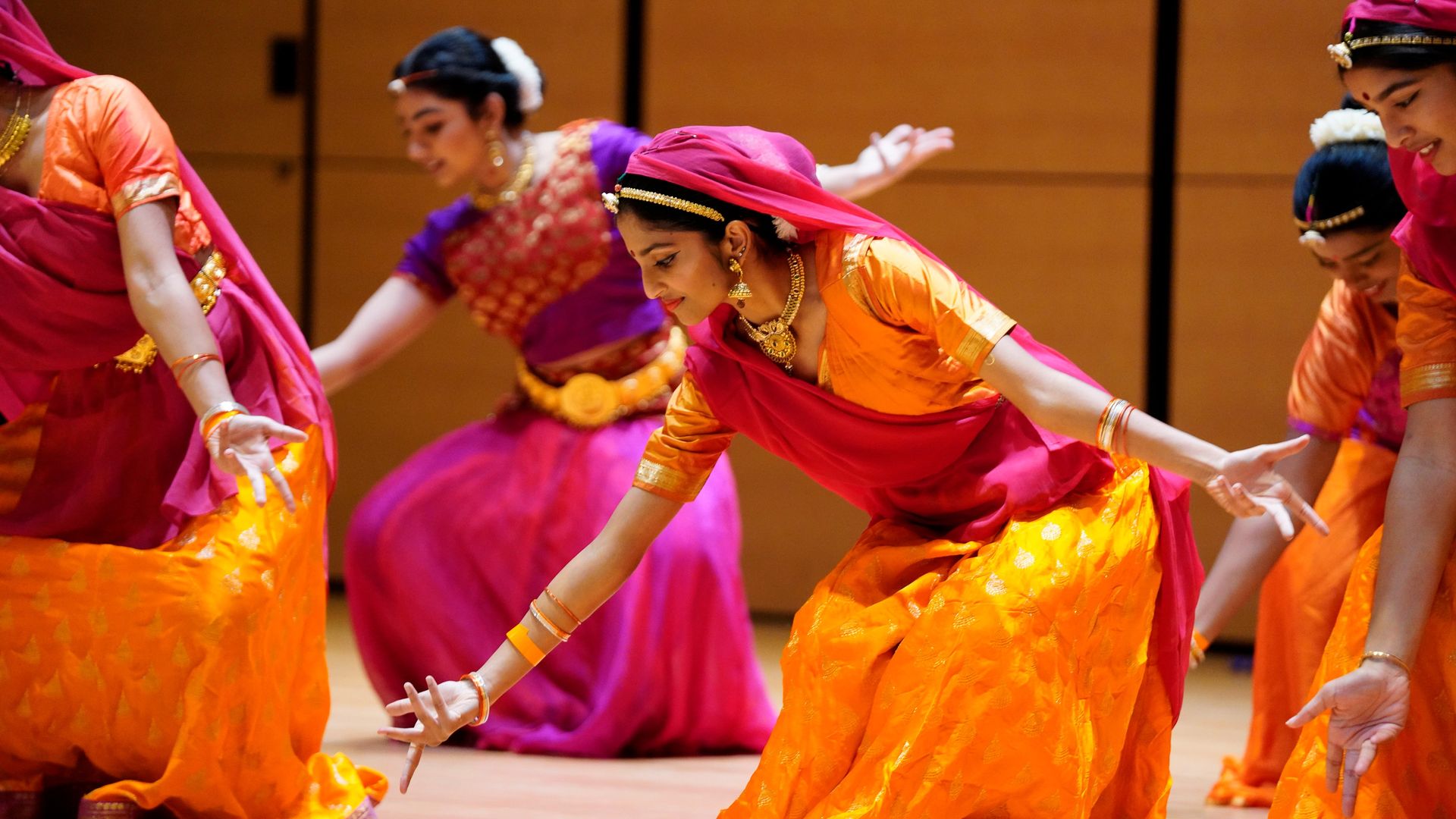 A group of women perform a dance wearing vibrant orange, pink, and purple outfits with gold jewelry.