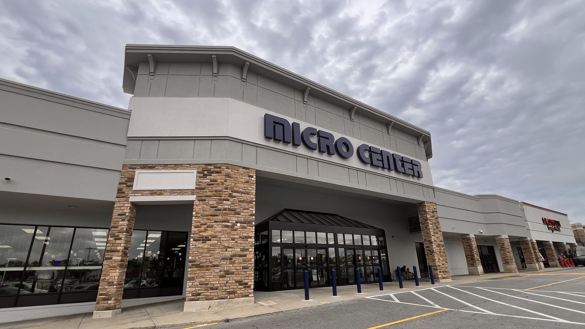 Wide view of a shopping center storefront with stone columns, glass windows, and a bold "MICRO CENTER" sign; gray, cloudy sky above and a yellow curb in the parking lot.