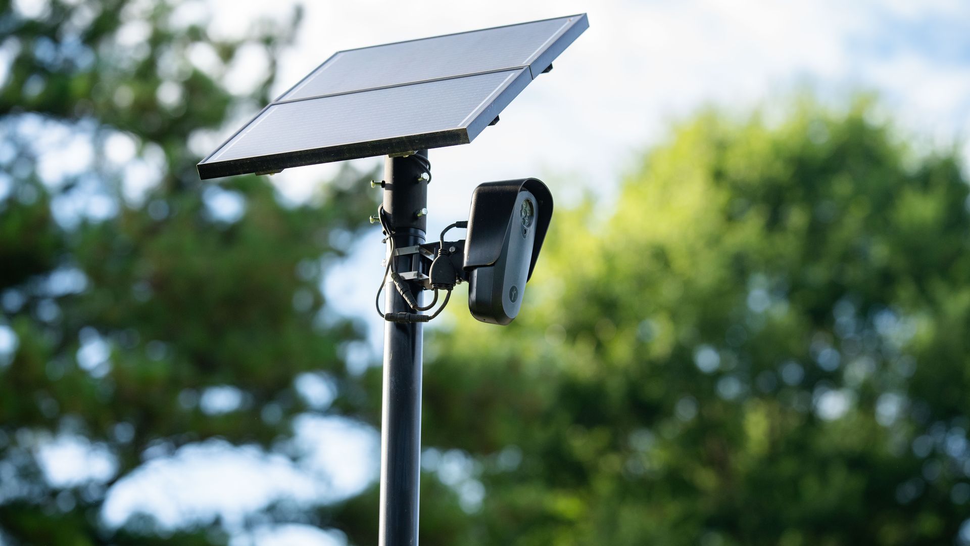 Solar panel array mounted on a pole with a license plate reading Flock camera housing beneath, outdoors with a blurred green treeline and blue sky in the background.