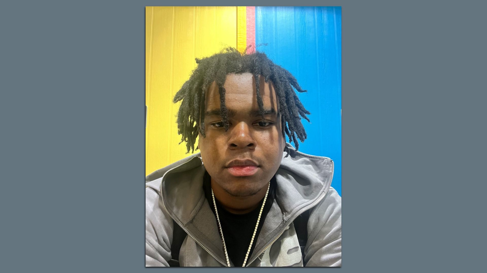 Close-up portrait of a young man with dreadlocks, wearing a gray jacket and gold chain, in front of a yellow, pink, and blue striped background.