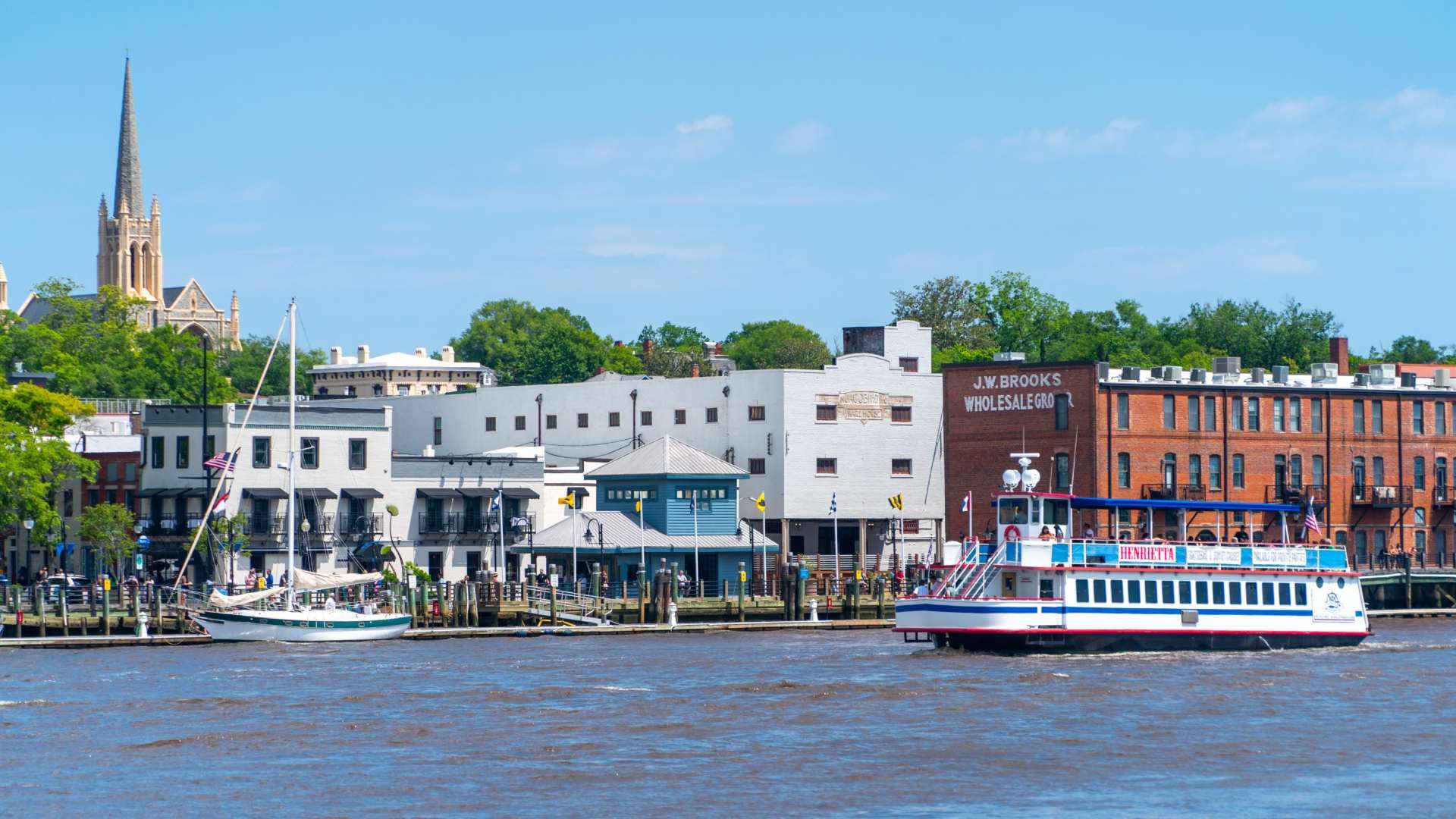 Sunny riverside town with a tall church steeple on the left, white and brick buildings along the waterfront, and a large white-and-blue passenger boat by the pier; a small sailboat nearby.