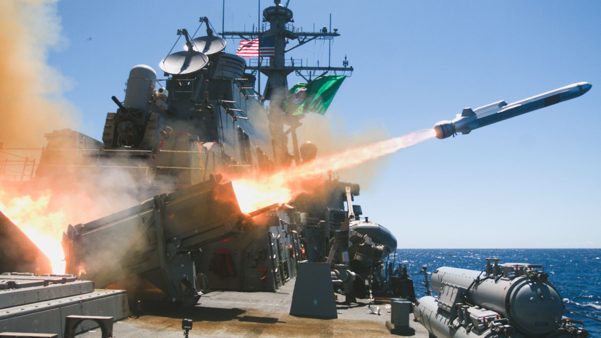 A naval missile rockets off the deck of a U.S. warship. Fire shoots out the back as it flies to the right. The ocean and sky behind the ship are different shades of blue,