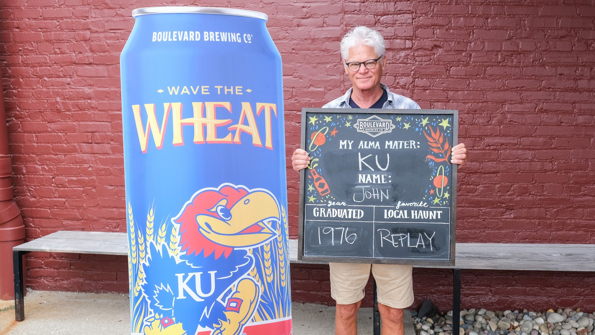 Older man with white hair and glasses holding a blackboard with KU alma mater, name John, graduated 1976, and favorite local haunt Replay, standing next to a large blue beer can prop against a red brick wall.