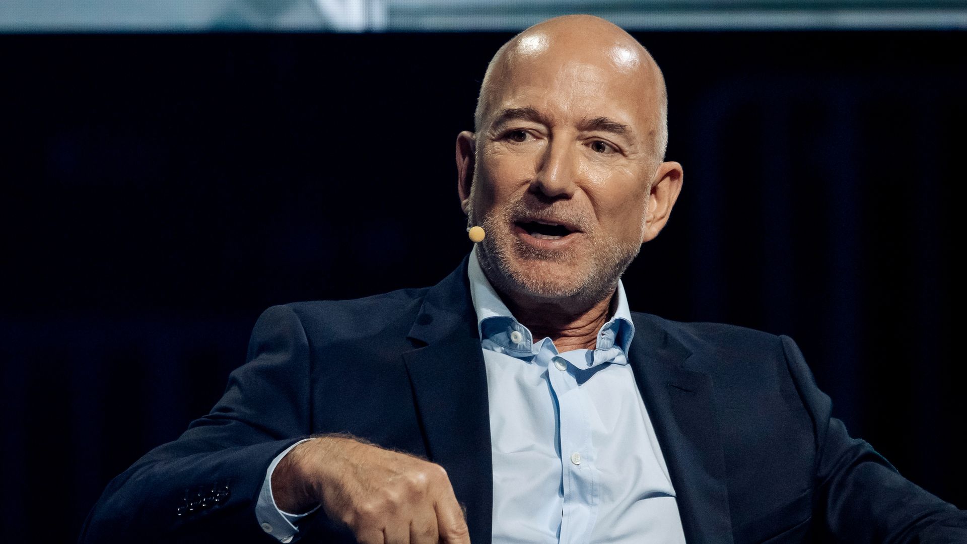 A person with a light beard wearing a dark blazer and light blue shirt, speaking with a headset mic against a dark background.