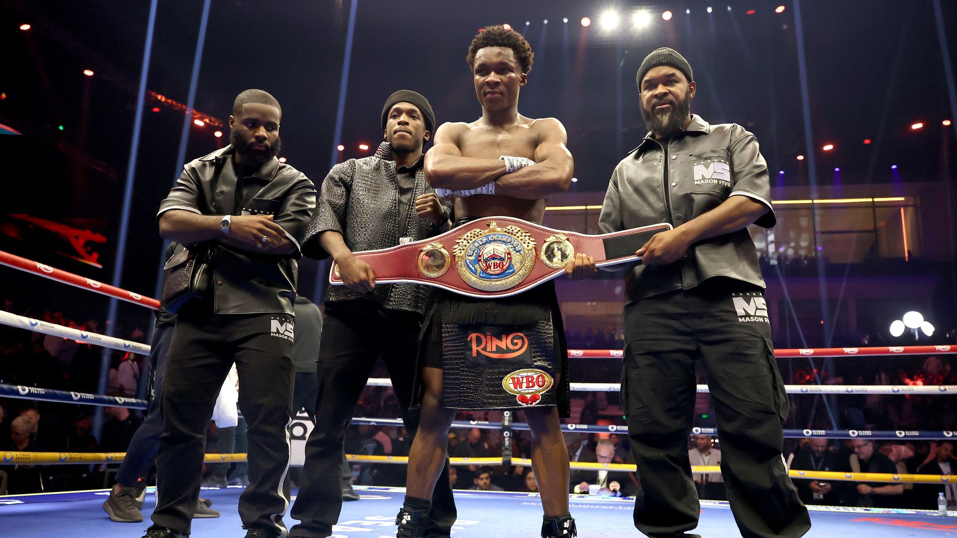 Four men stand in a boxing ring; center is a shirtless champion with a large belt. Flanked by two helpers and a man in gray, bright arena lights and ropes surround them.