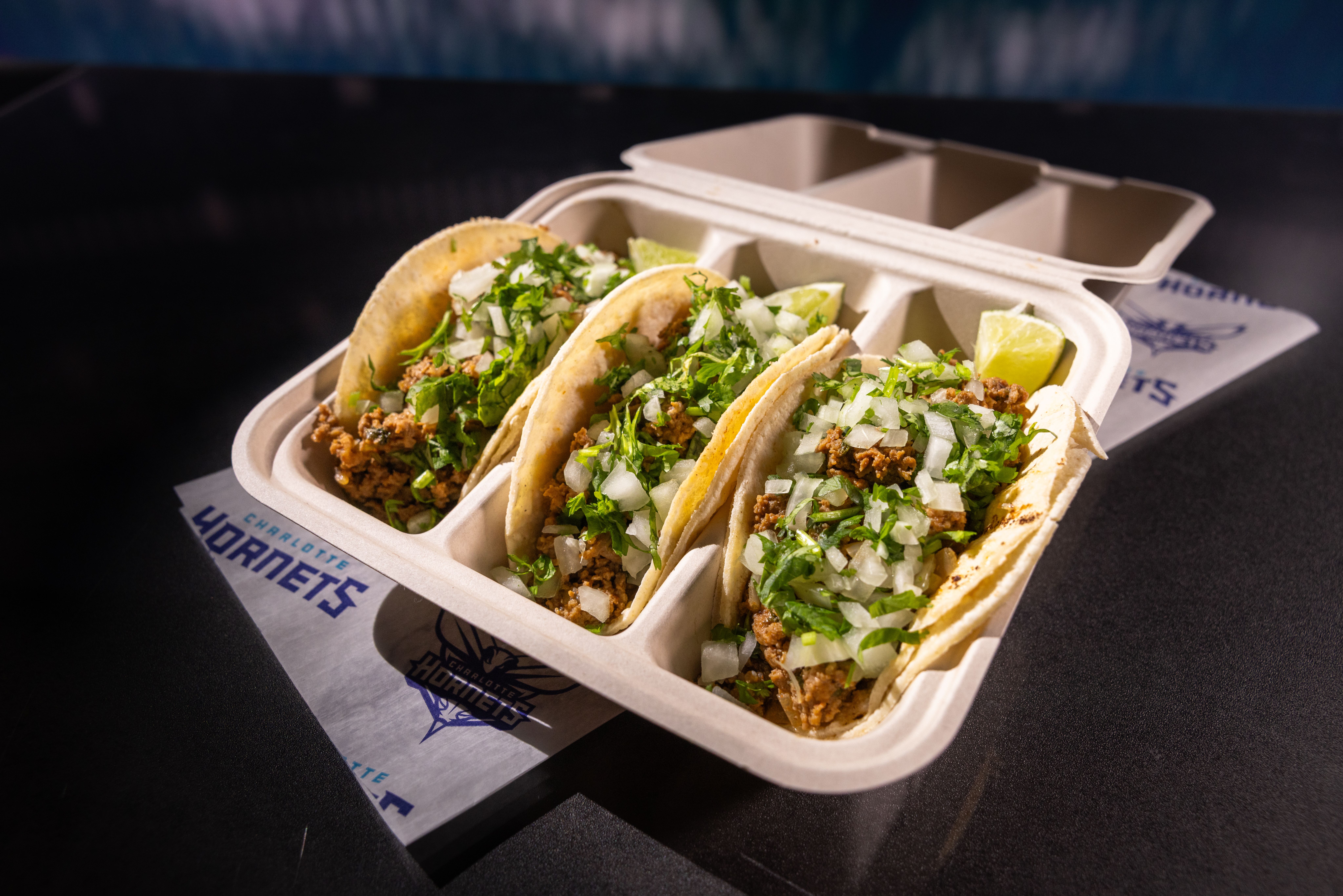 Three tacos with ground meat, chopped onions, and cilantro in a beige divided takeout container with lime wedges, on a table with a Charlotte Hornets napkin underneath.
