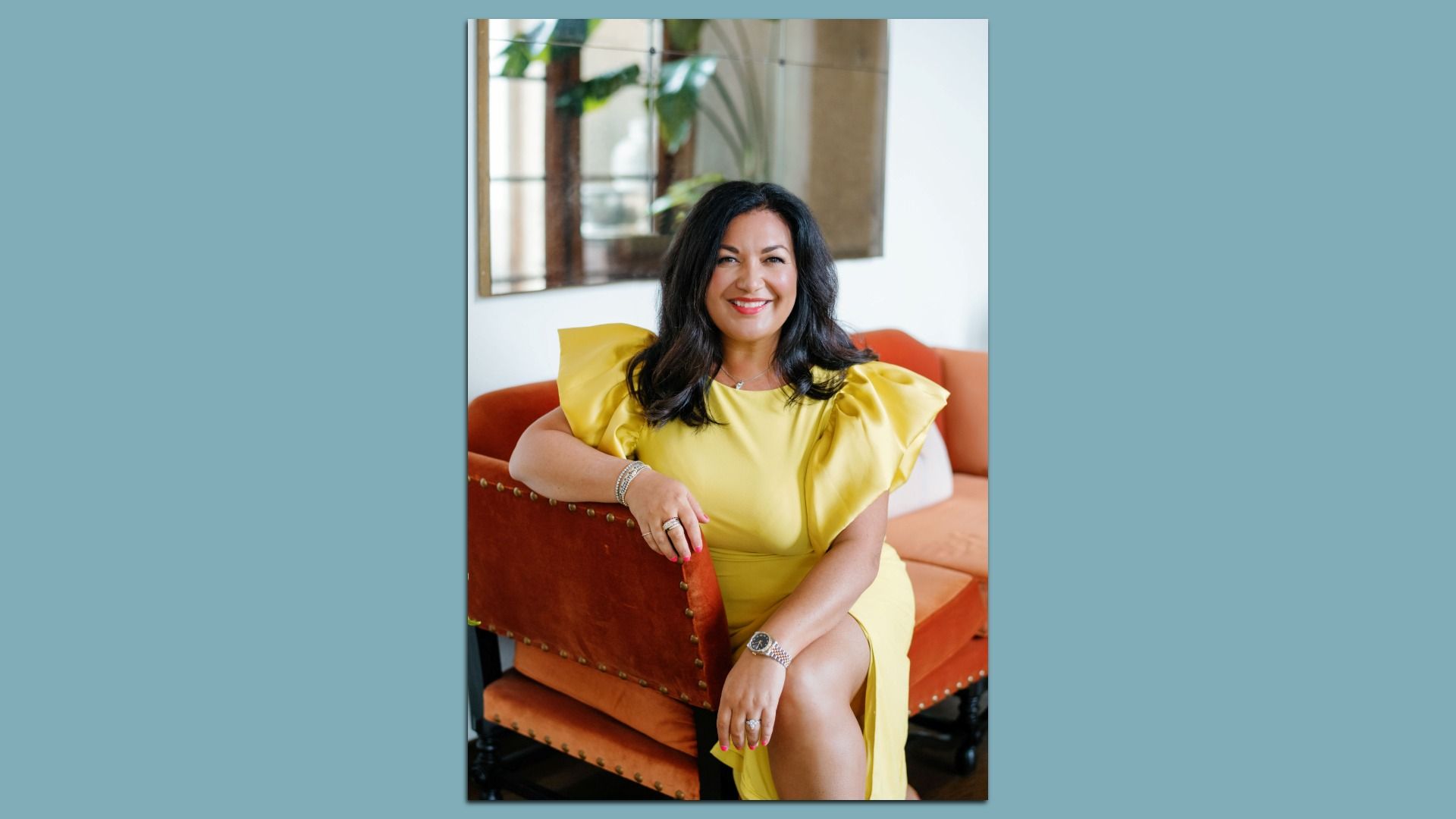 Woman with black hair in a yellow dress with ruffled sleeves sitting on an orange couch with brass studs, smiling, wearing bracelets and a watch, with a mirror and plants in the background.