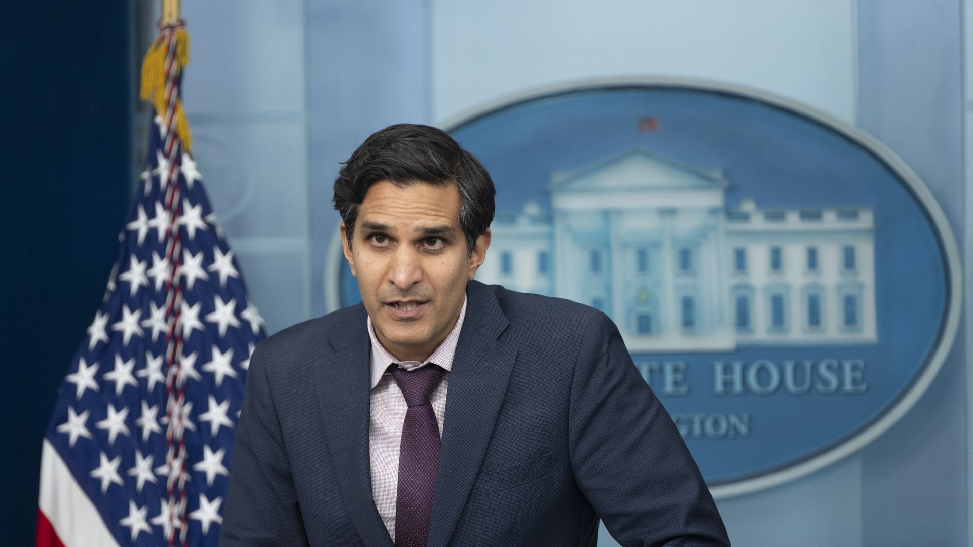 A middle-age male with a blue suit leans over the White House lectern in a press briefing
