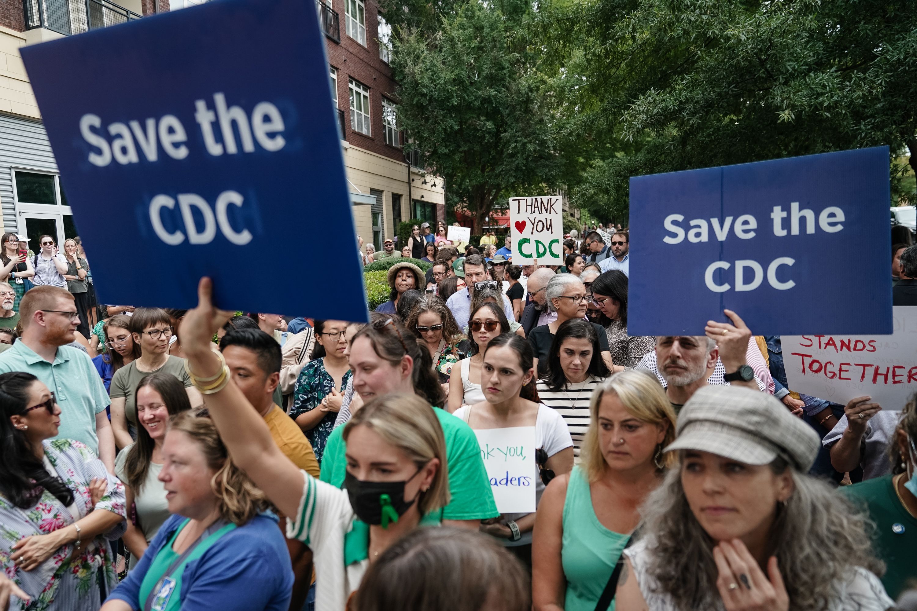 Crowd of diverse people at a protest holding blue signs reading "Save the CDC" and other signs thanking the CDC, gathered outdoors near trees and buildings.