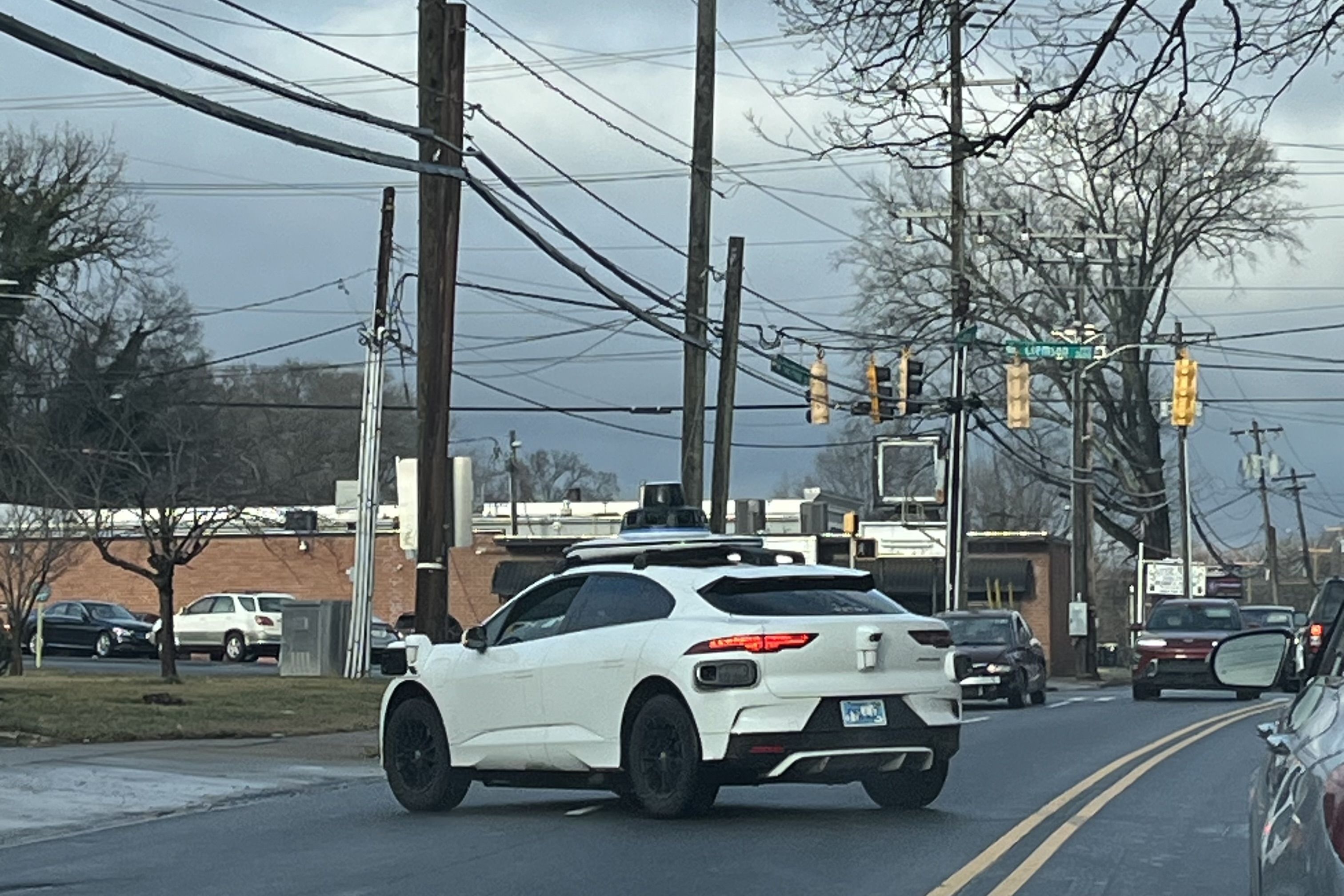 White autonomous car with roof sensors turning left on a street under a cloudy sky, with traffic lights and a brick building in the background.