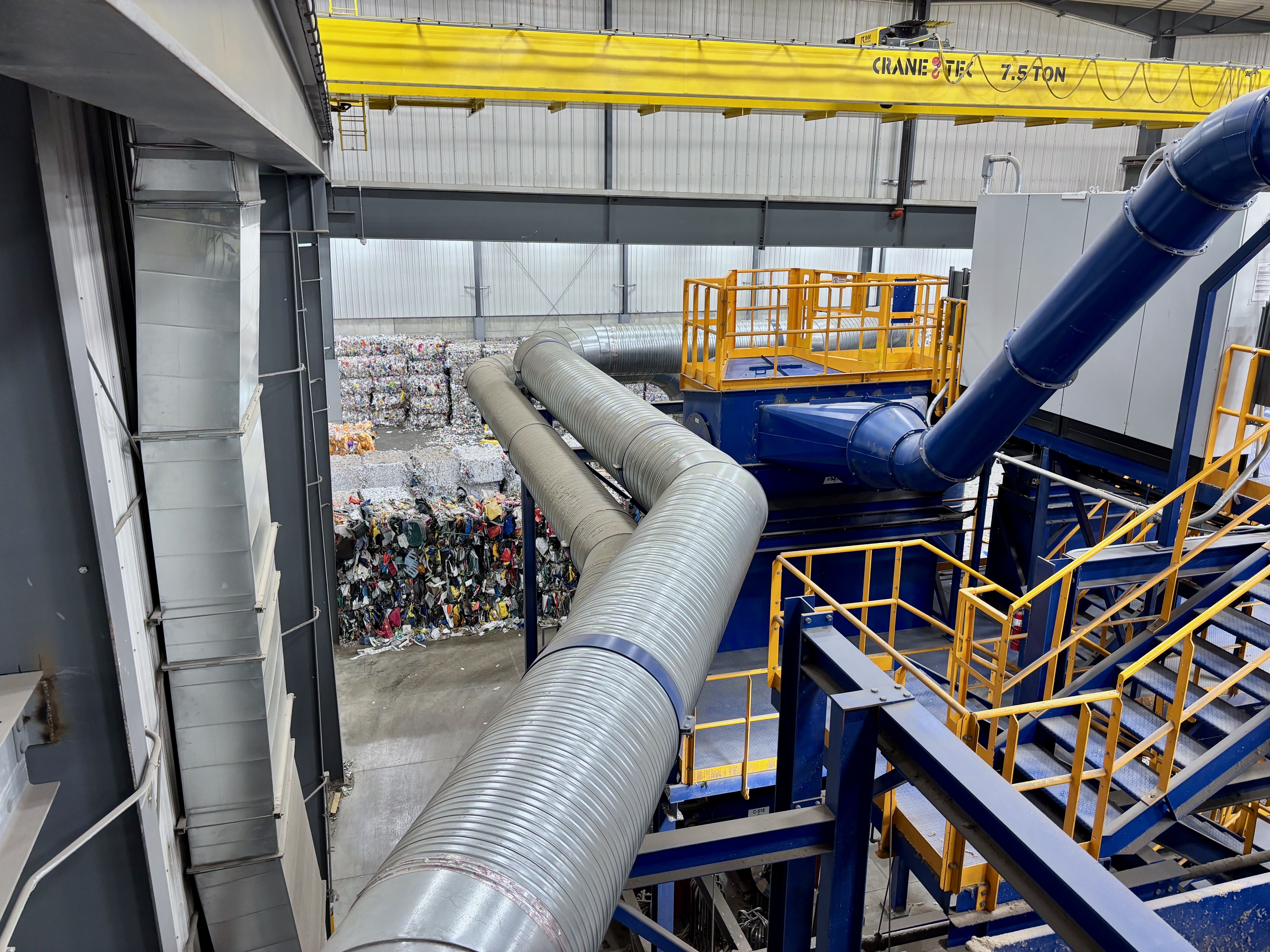 Industrial interior with large galvanized ductwork, a bright yellow overhead crane labeled 7.5 TON, blue and yellow railings, gray metal walls, and a pile of compacted waste outside.