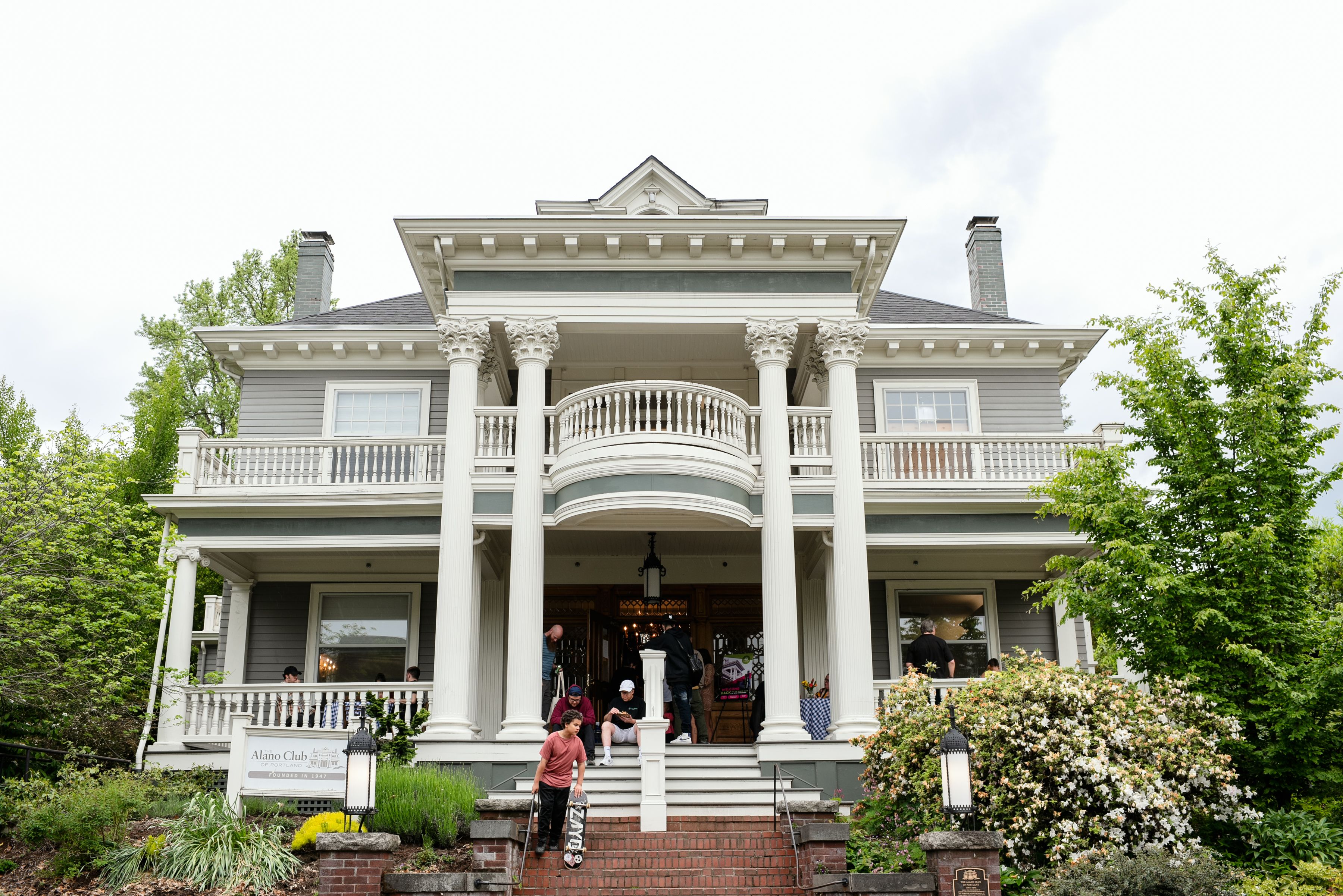Large historic gray and white two-story house with columns and balcony, people on the steps and porch, green trees and bushes around, sign reads Alano Club of Portland.