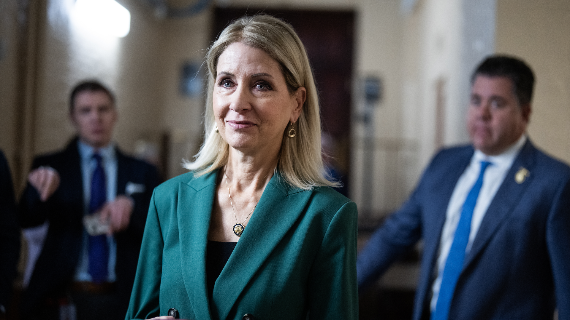 Rep. Mary Miller, wearing a turquoise suit and holding a phone, walks through a white brick hallway along with reporters and other members of Congress.