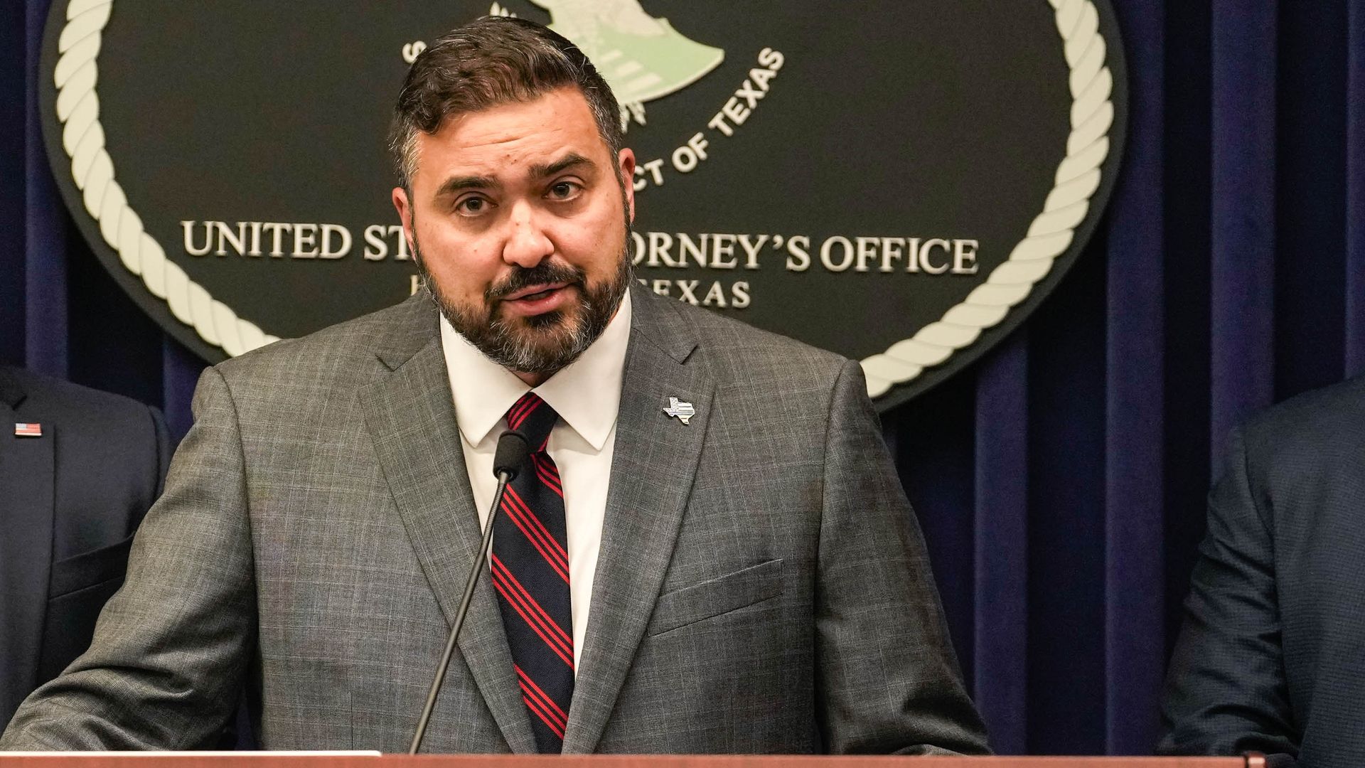 A dark-haired, bearded U.S. Attorney Nicholas Ganjei, wearing a gray suit, white shirt and red and blue striped tie, speaks at at a DOJ podium.