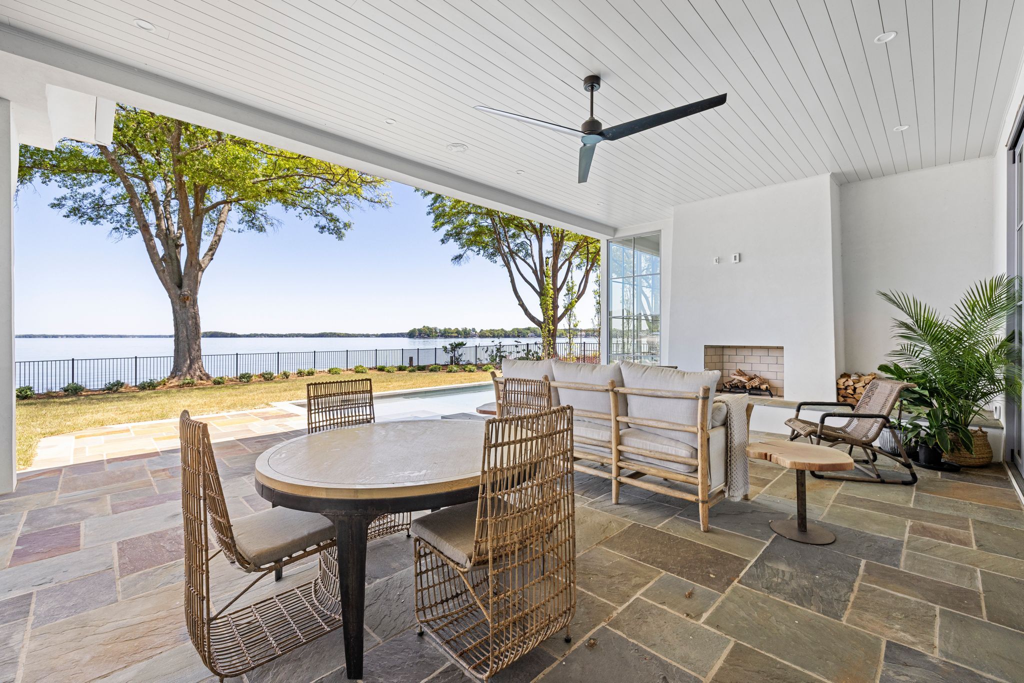 Covered patio with natural stone floor, wicker dining chairs around round table, cushioned sofa, fireplace, ceiling fan, plants, overlooking lake and trees under clear sky.