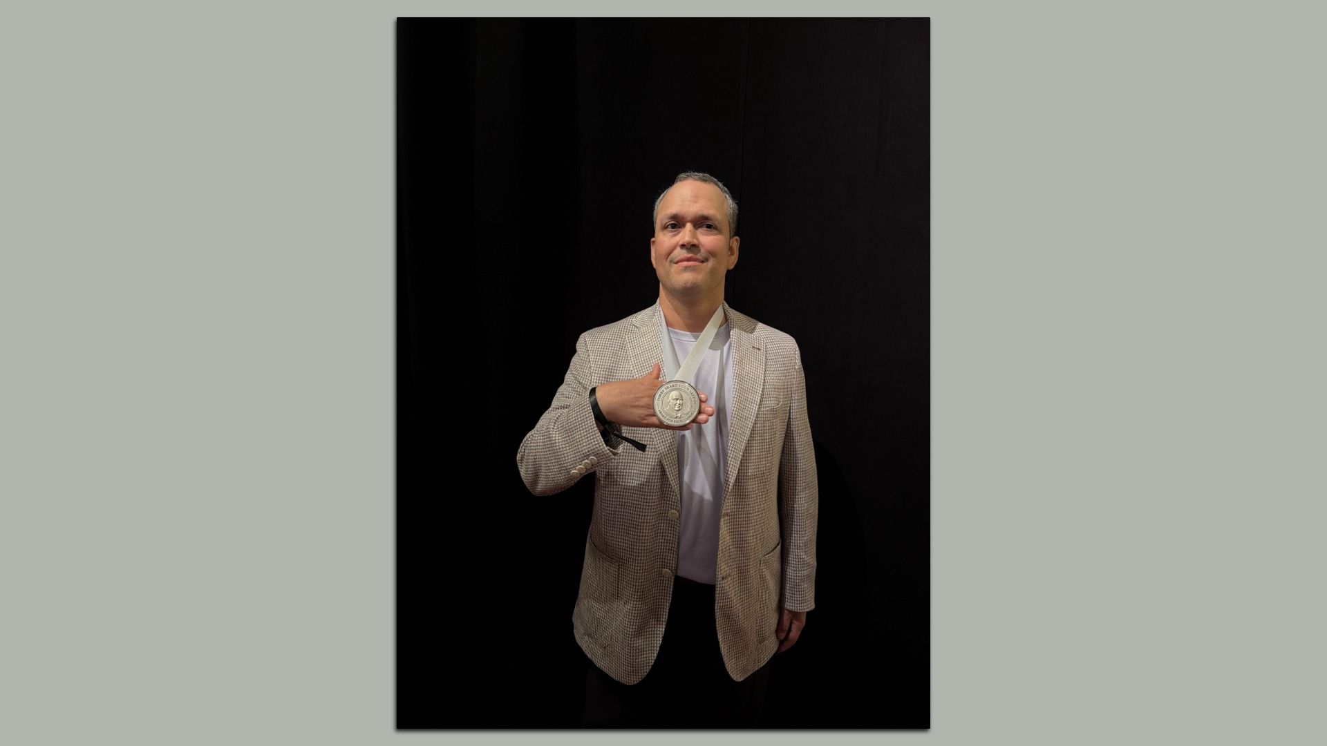 A man in a beige checkered blazer holds a silver medal on a white ribbon, smiling slightly, against a dark backdrop.