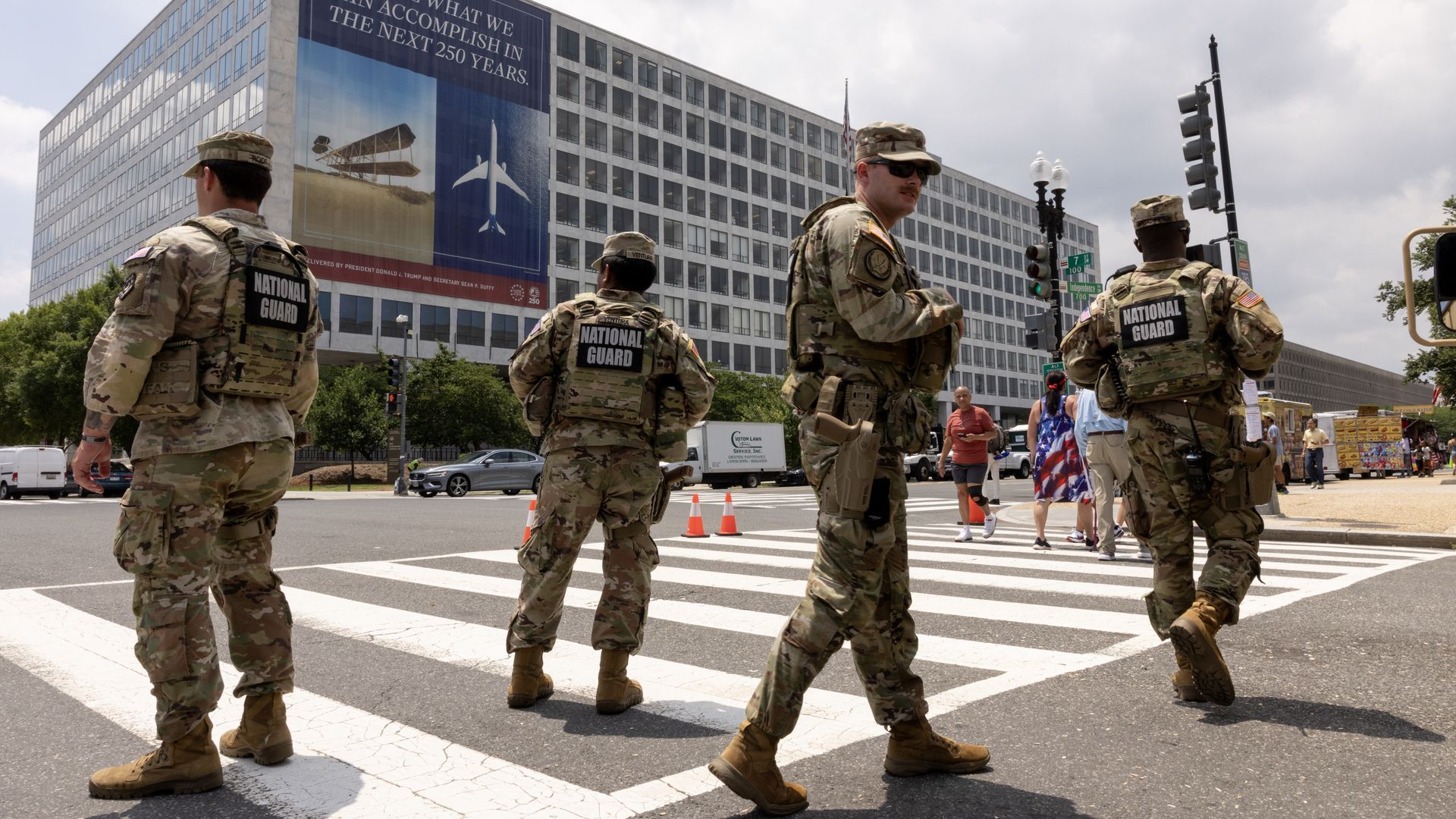 National Guard soldiers in camouflage stand at a crosswalk in front of a large office building with a banner reading, "Imagine what we can accomplish in the next 250 years."
