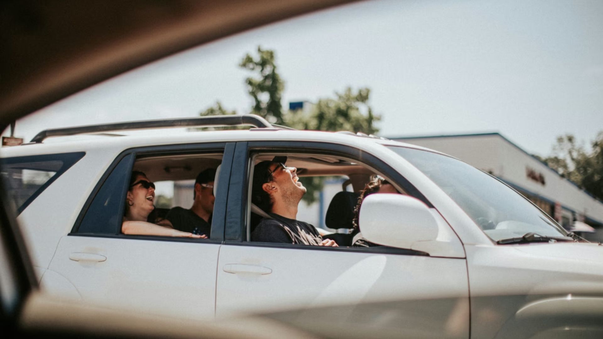 A white SUV with three passengers laughing inside; a man in the front seat wears sunglasses and tilts his head back, while others smile. Sunny day with trees and storefronts in the background.