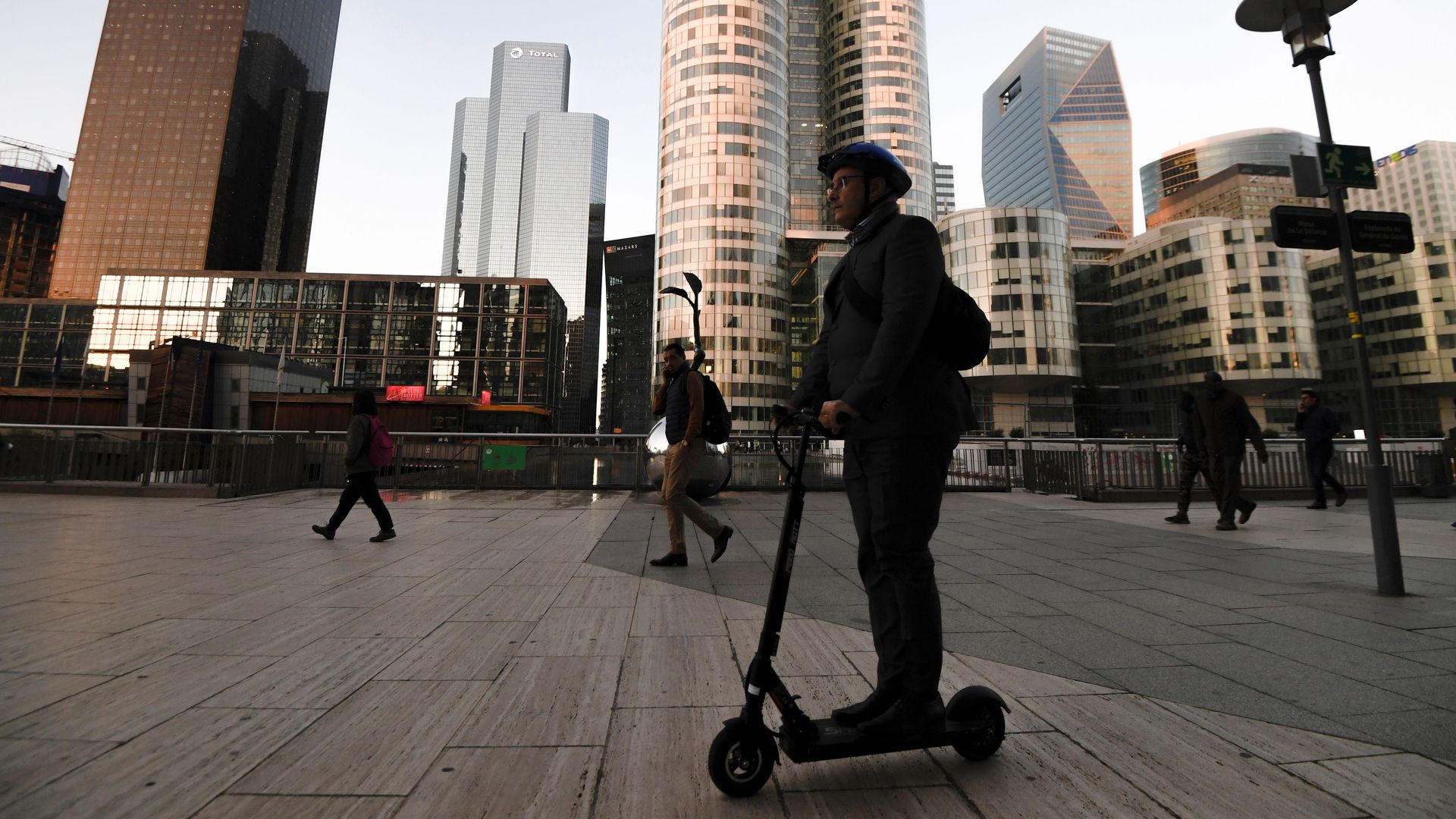 In this image, a person rides an electric scooter through a mostly empty city quad. Skyscrapers are in the background.