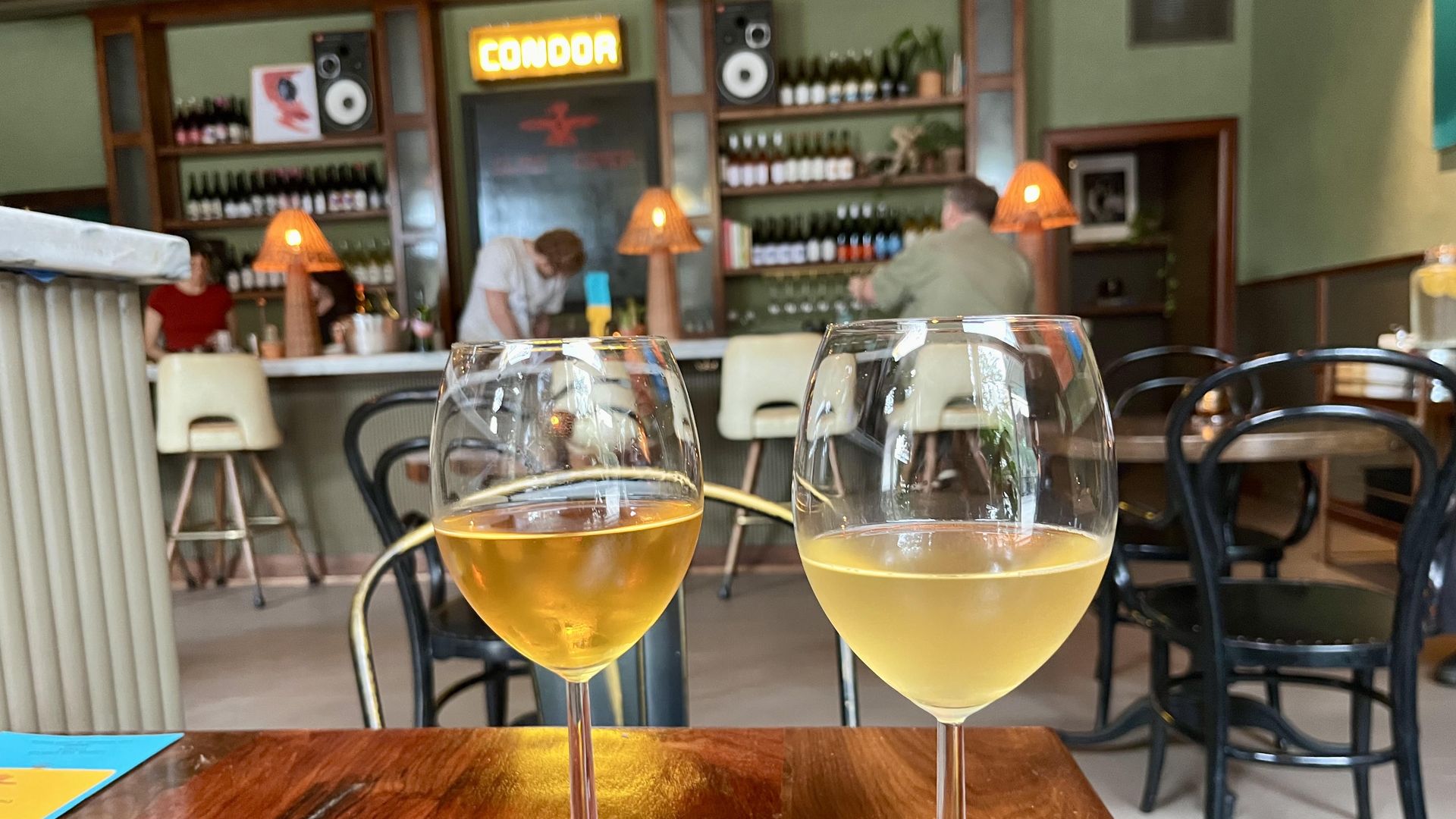 Two wine glasses on a wooden table in a cozy bar, with blurred background shelves of bottles, glowing orange lamps, and a glowing "CONDOR" sign at Condor Wine Bar in San Antonio.