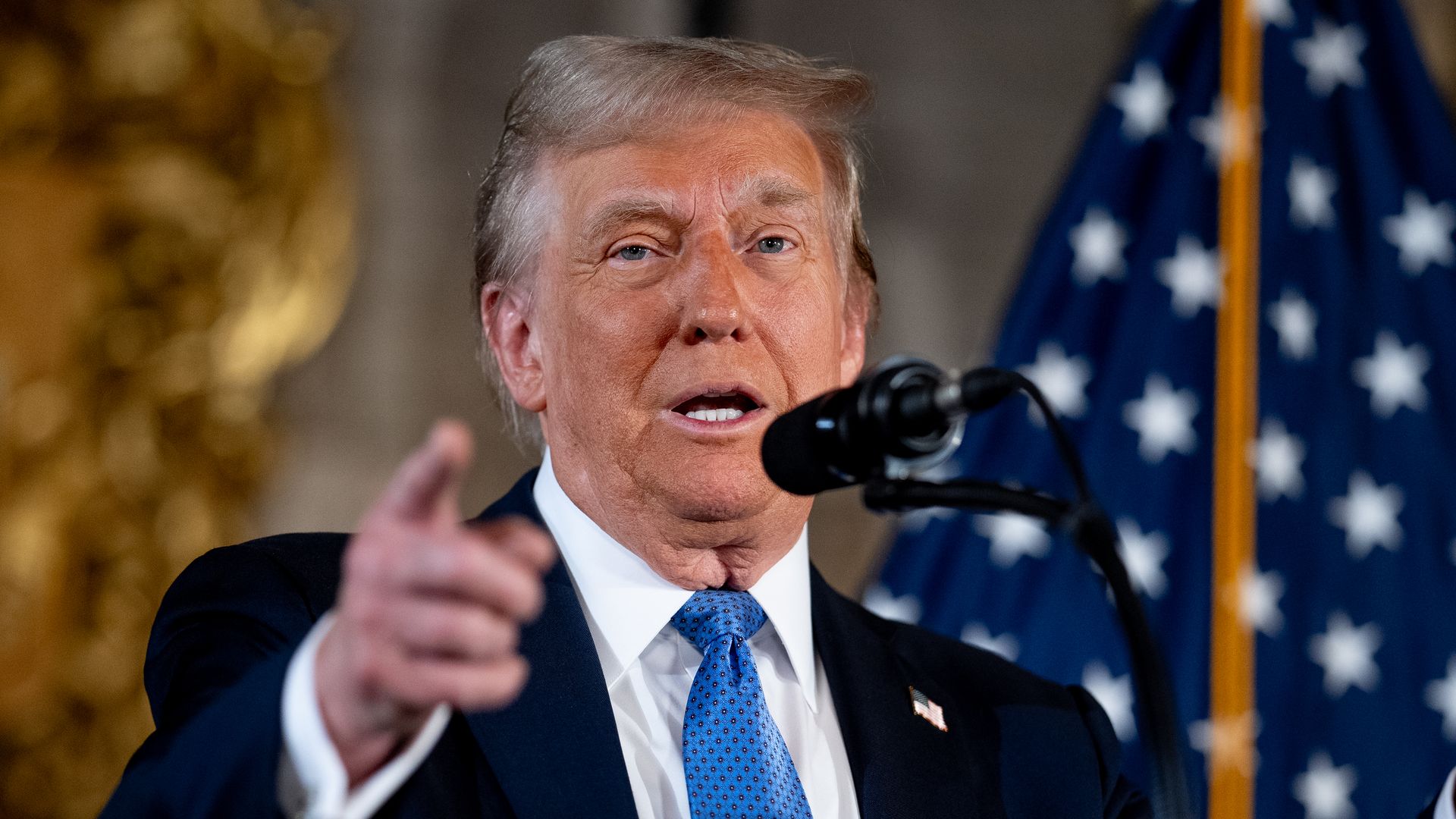 Donald Trump, wearing a dark suit and blue tie, speaks into a microphone. One hand is pointing in the direction of the camera. 