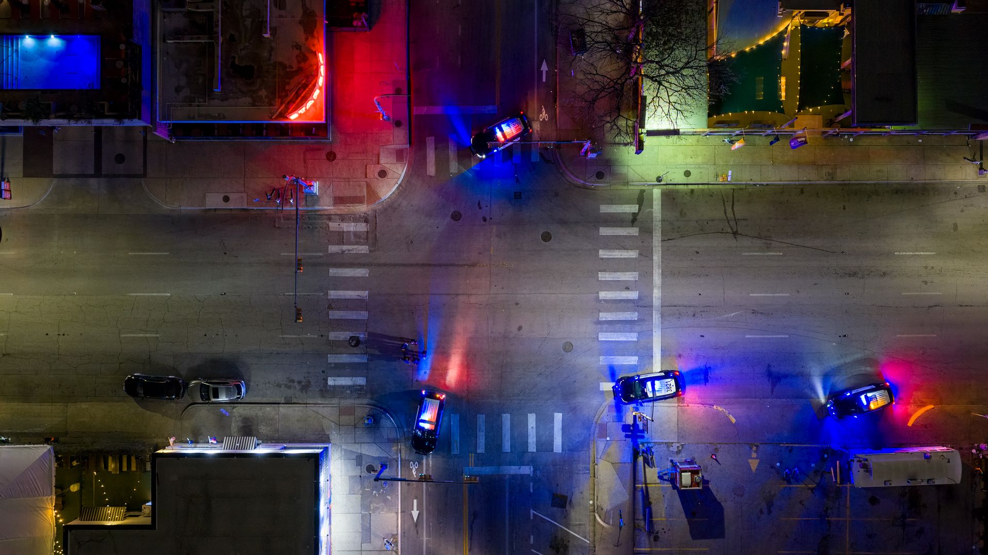 Aerial night view of a street intersection with four police cars flashing red and blue lights, casting colorful glows on the pavement and nearby buildings.