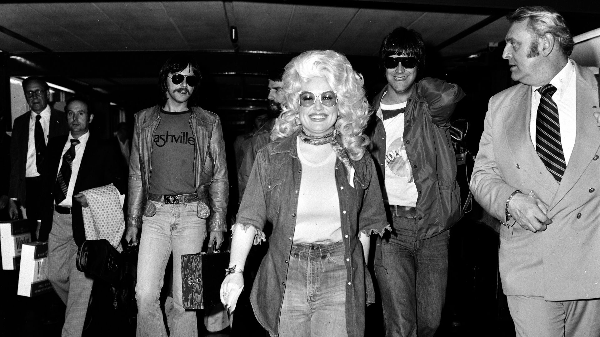 Dolly Parton walking through a crowded airport smiling in a black and white photograph from 1977.